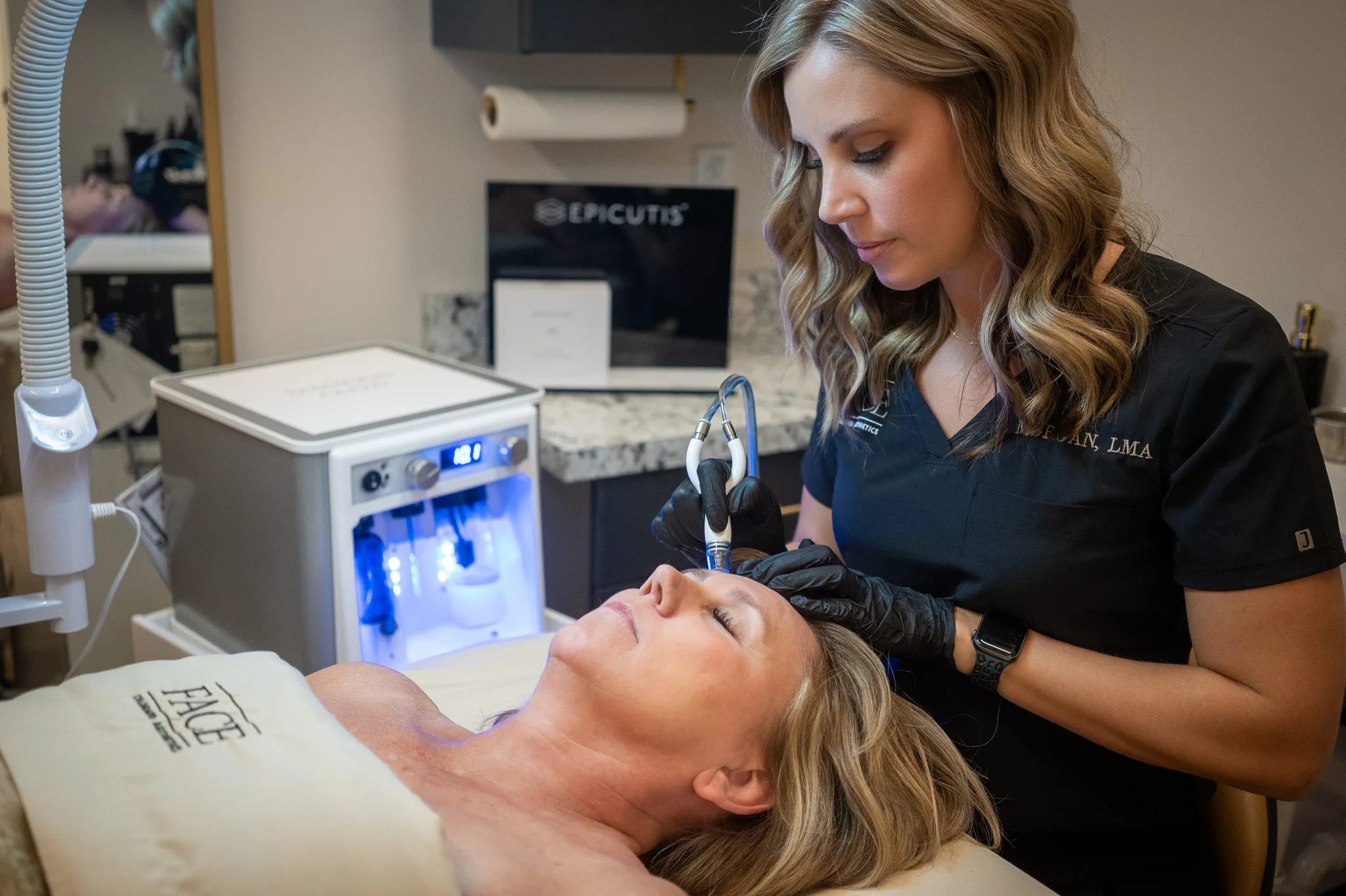A FACE Colorado provider in a black scrub top performs a facial treatment on a reclined client using a handheld device linked to a machine.