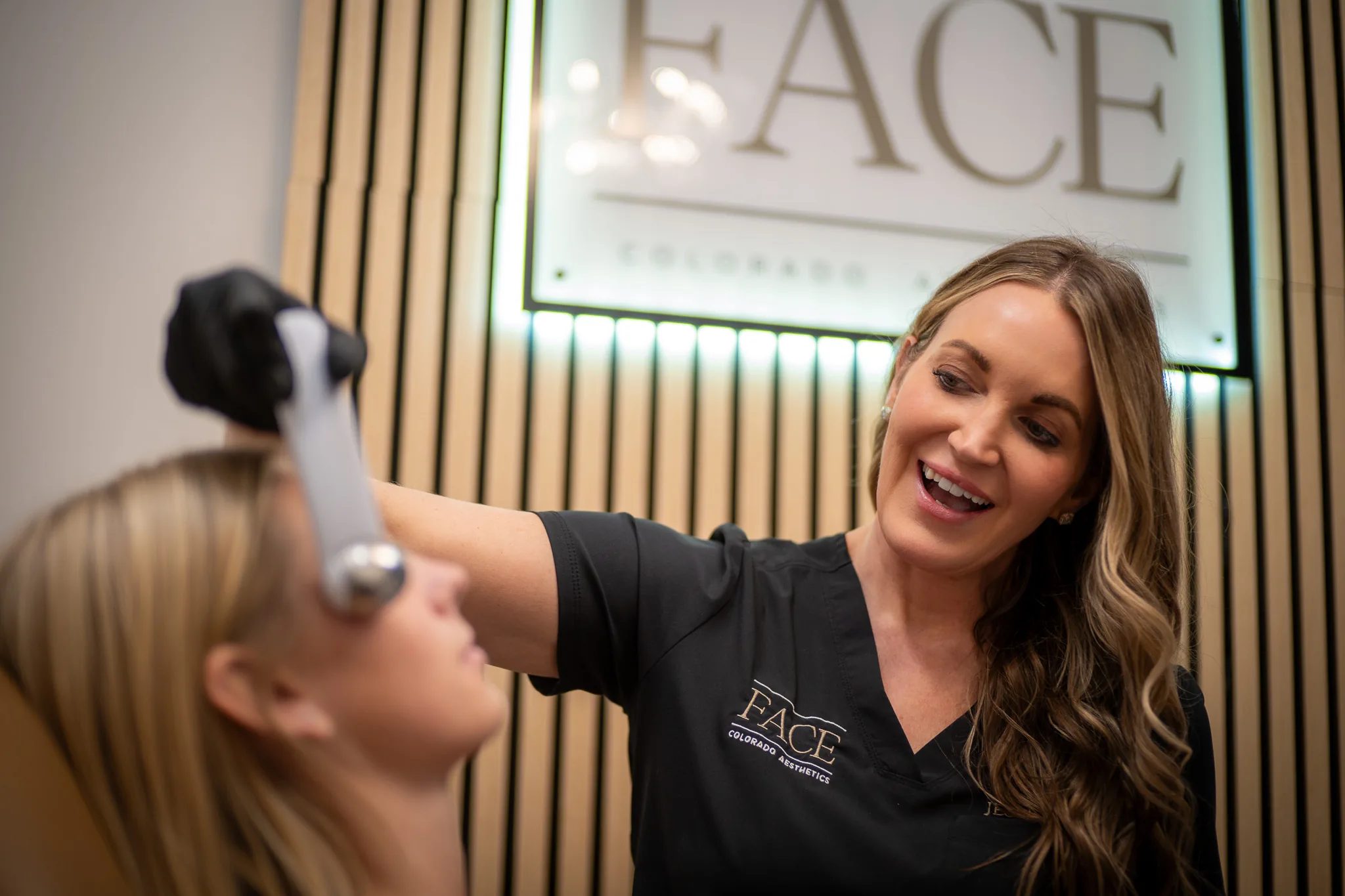 A laughing FACE Colorado nurse in a black scrub top holds a metal facial globe to a client's forehead beneath the illuminated FACE sign.