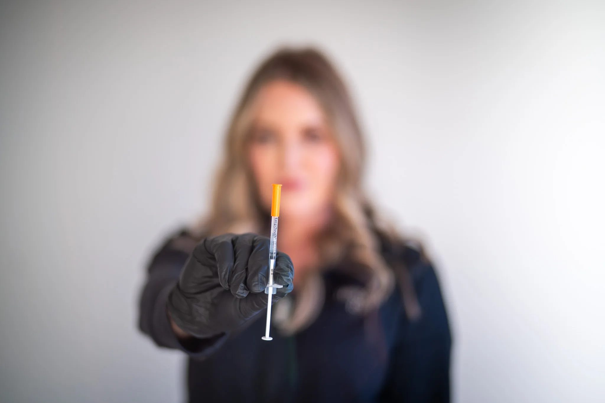 A black-gloved hand holds a small orange-capped syringe in sharp focus while a FACE Colorado nurse stands out of focus behind it.