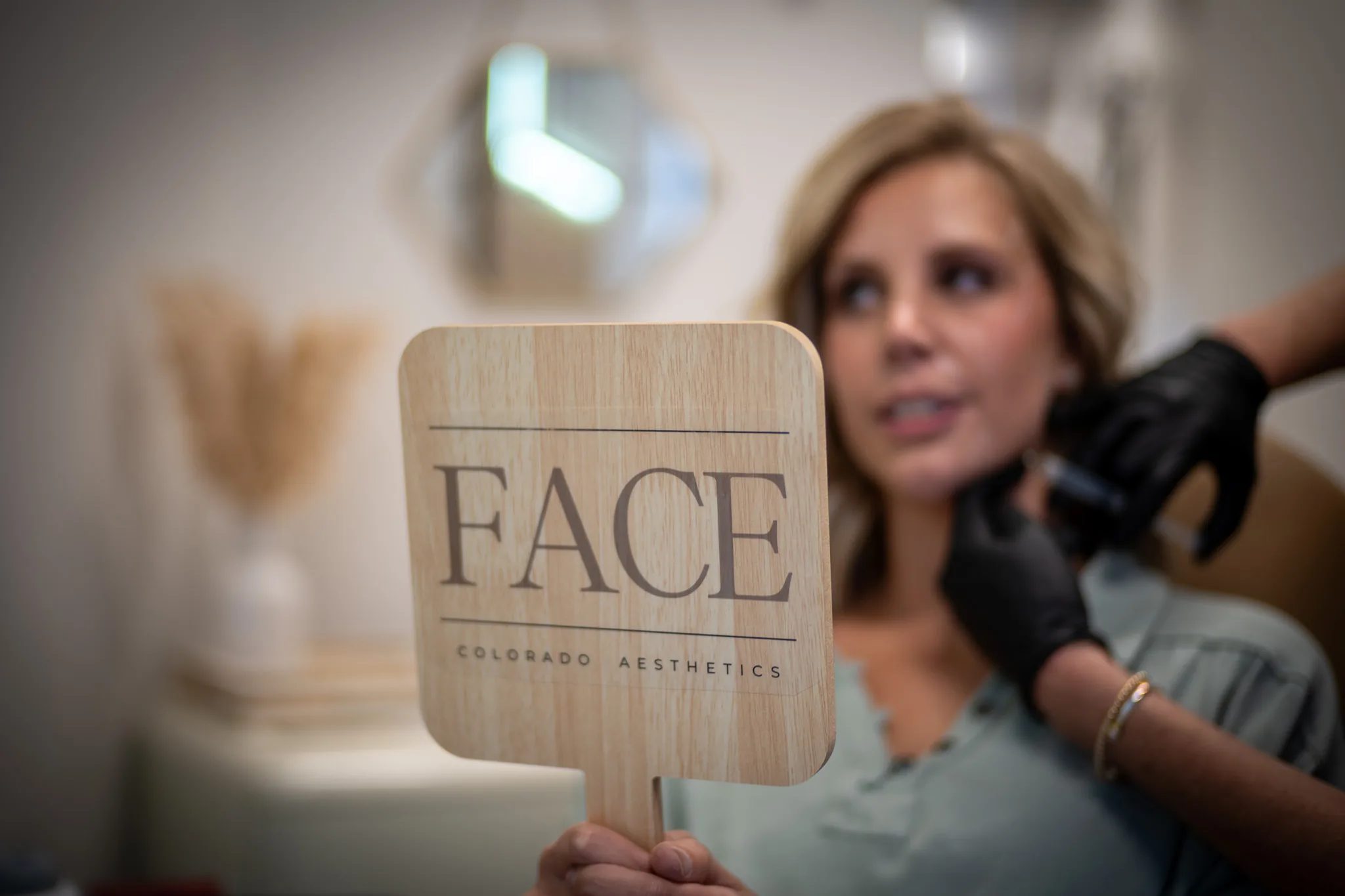 A wooden hand mirror printed with the FACE Colorado Aesthetics logo is held up in focus while a client receives treatment behind it.