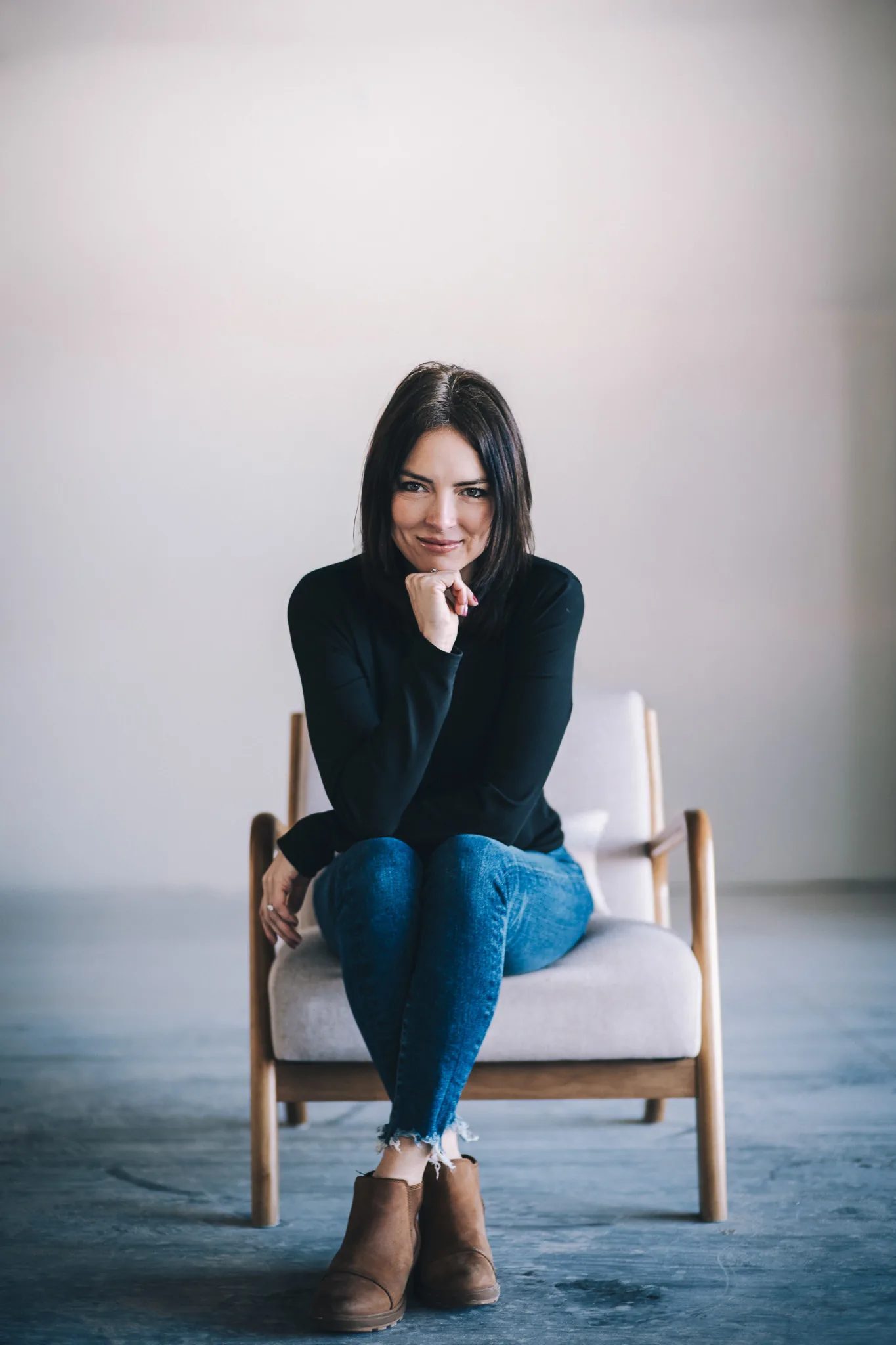 A FACE Colorado team member in a black top and jeans sits in a wooden armchair with chin resting on one hand.