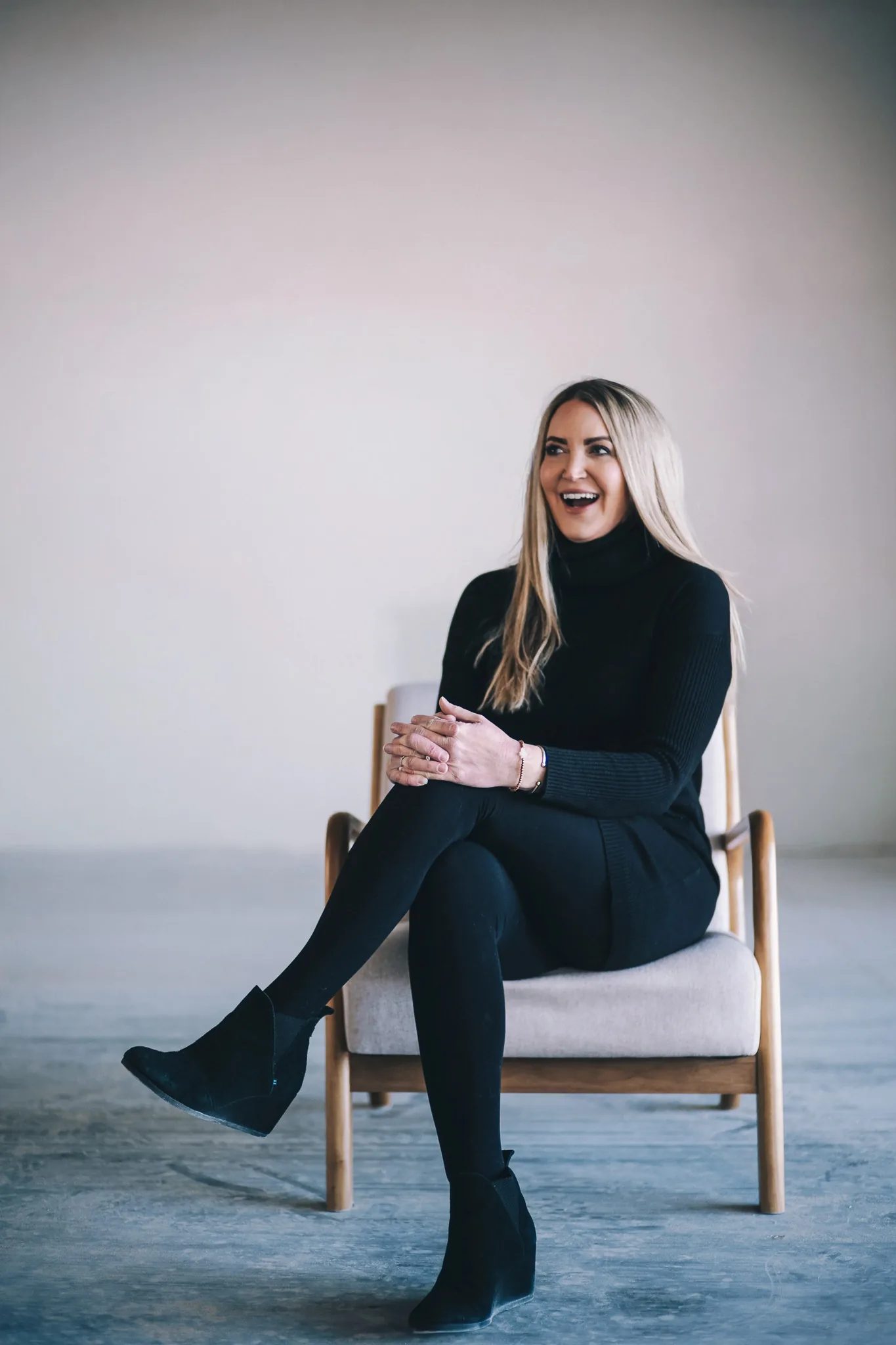 A FACE Colorado team member in a black turtleneck laughs while seated with legs crossed in a wooden armchair.