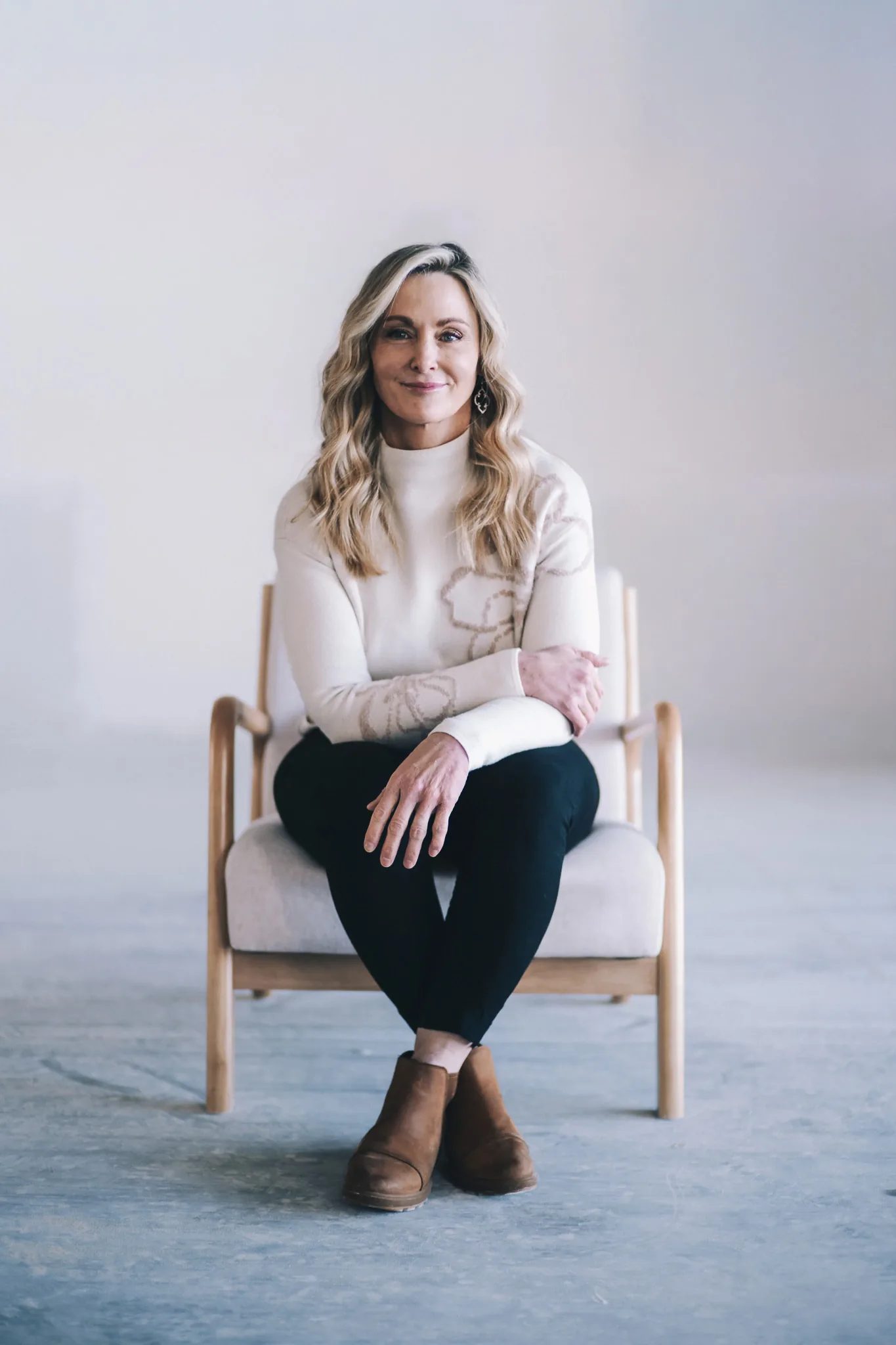 A FACE Colorado team member in a cream floral-motif sweater sits in a wooden armchair with arms loosely crossed.