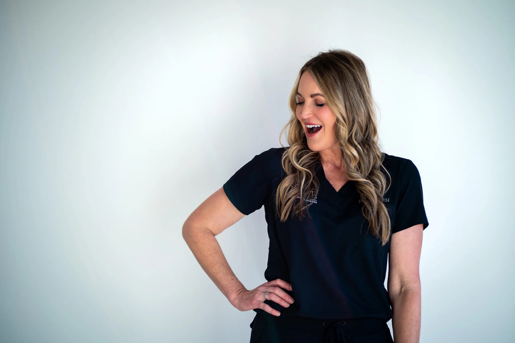 A FACE Colorado nurse in a black scrub top laughs with a hand on one hip against a bright white studio backdrop.