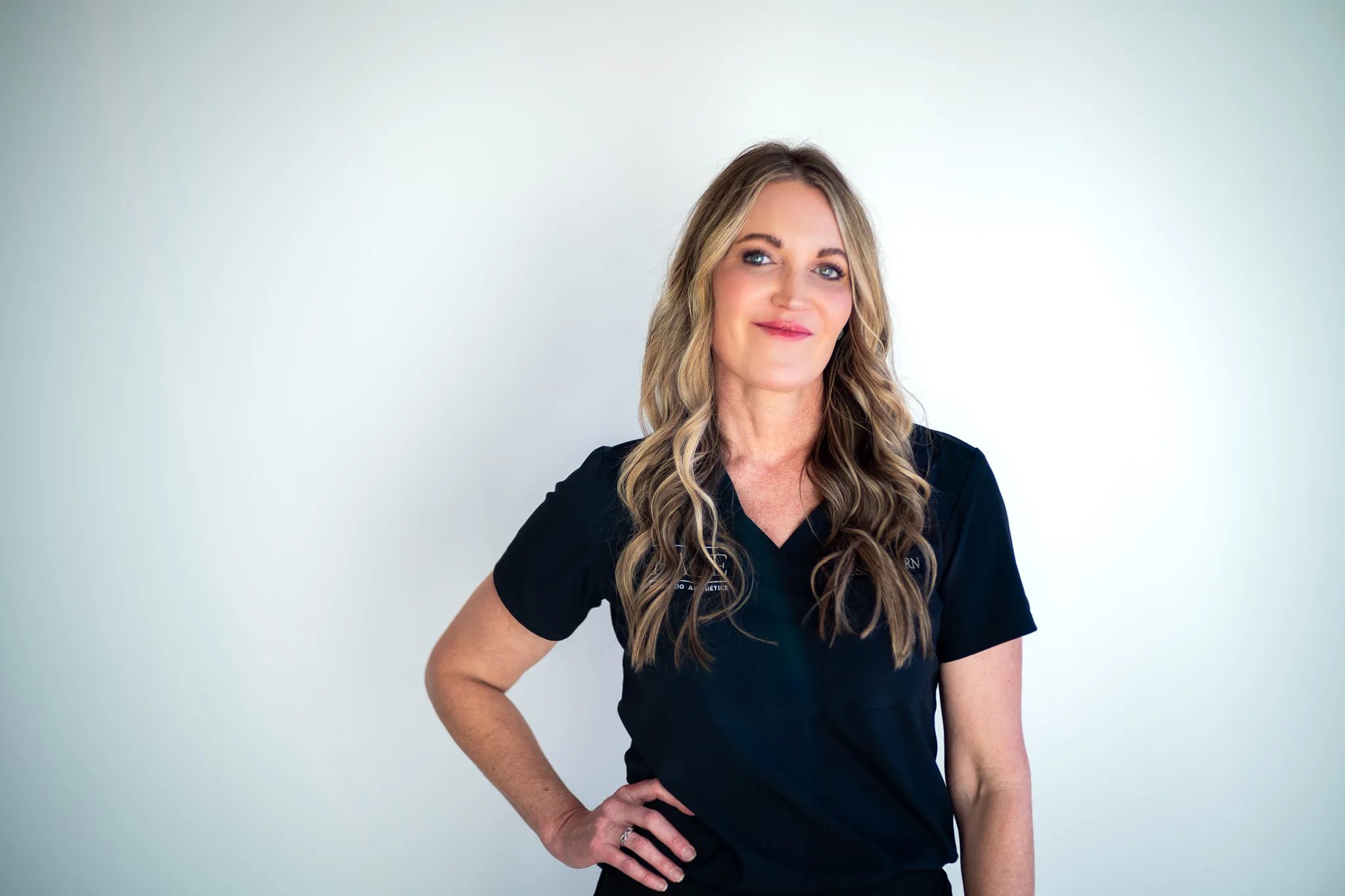 A FACE Colorado nurse in a black scrub top smiles at the camera with a hand on one hip in front of a white studio backdrop.
