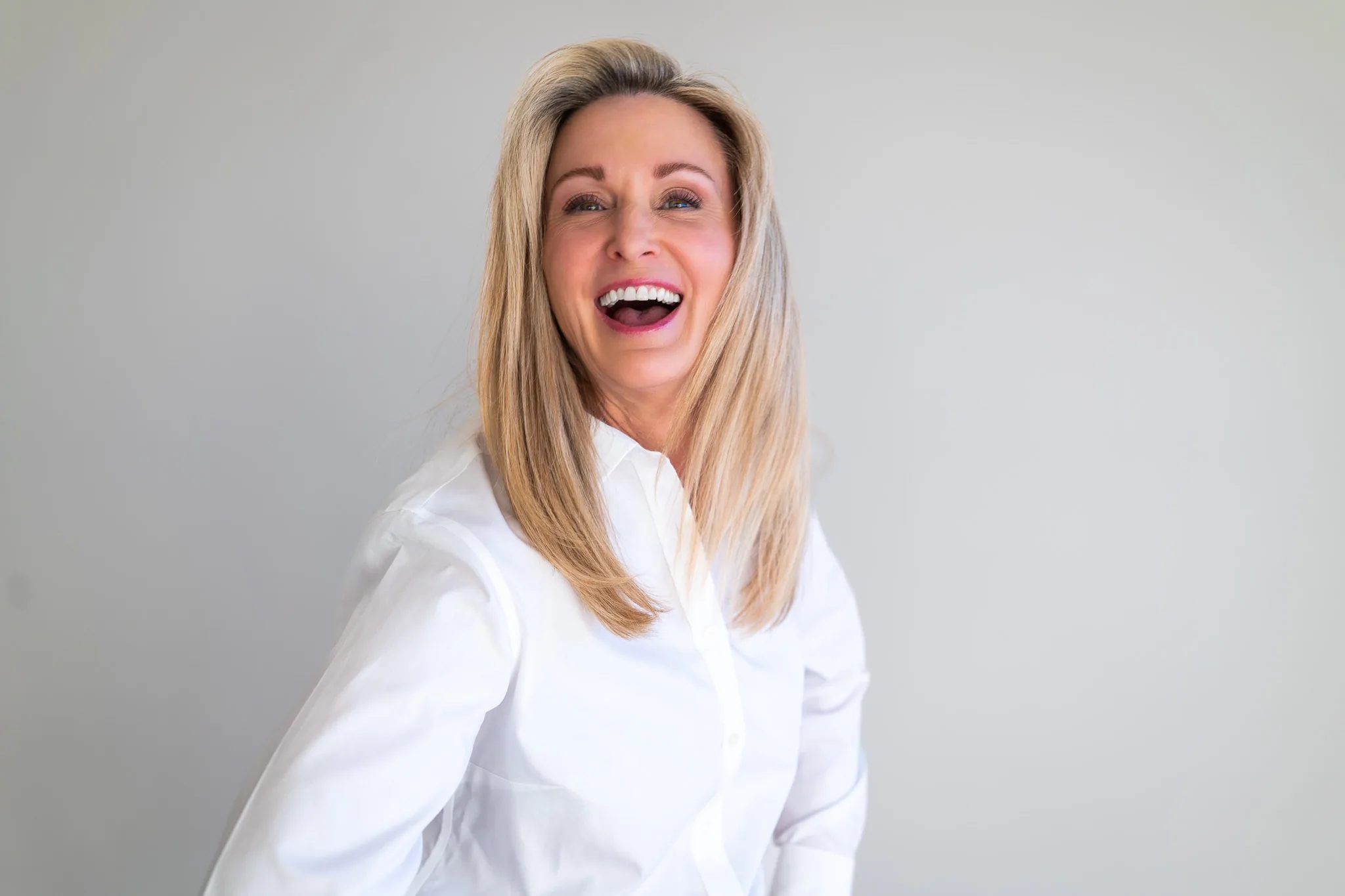 A FACE Colorado nurse in a crisp white button-up shirt laughs with head tilted back against a grey studio backdrop.