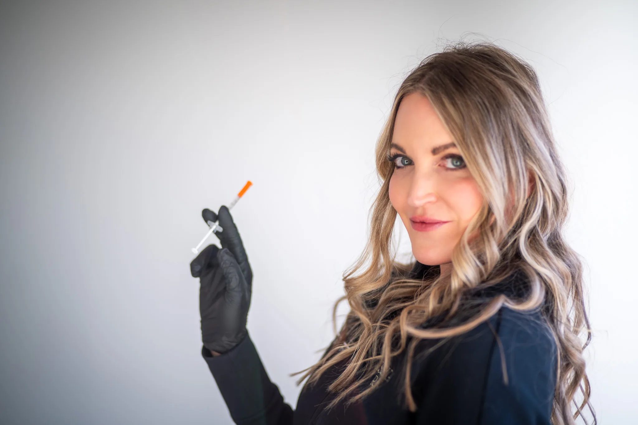 A FACE Colorado nurse in a black jacket and black glove holds up a small syringe while glancing back over one shoulder.