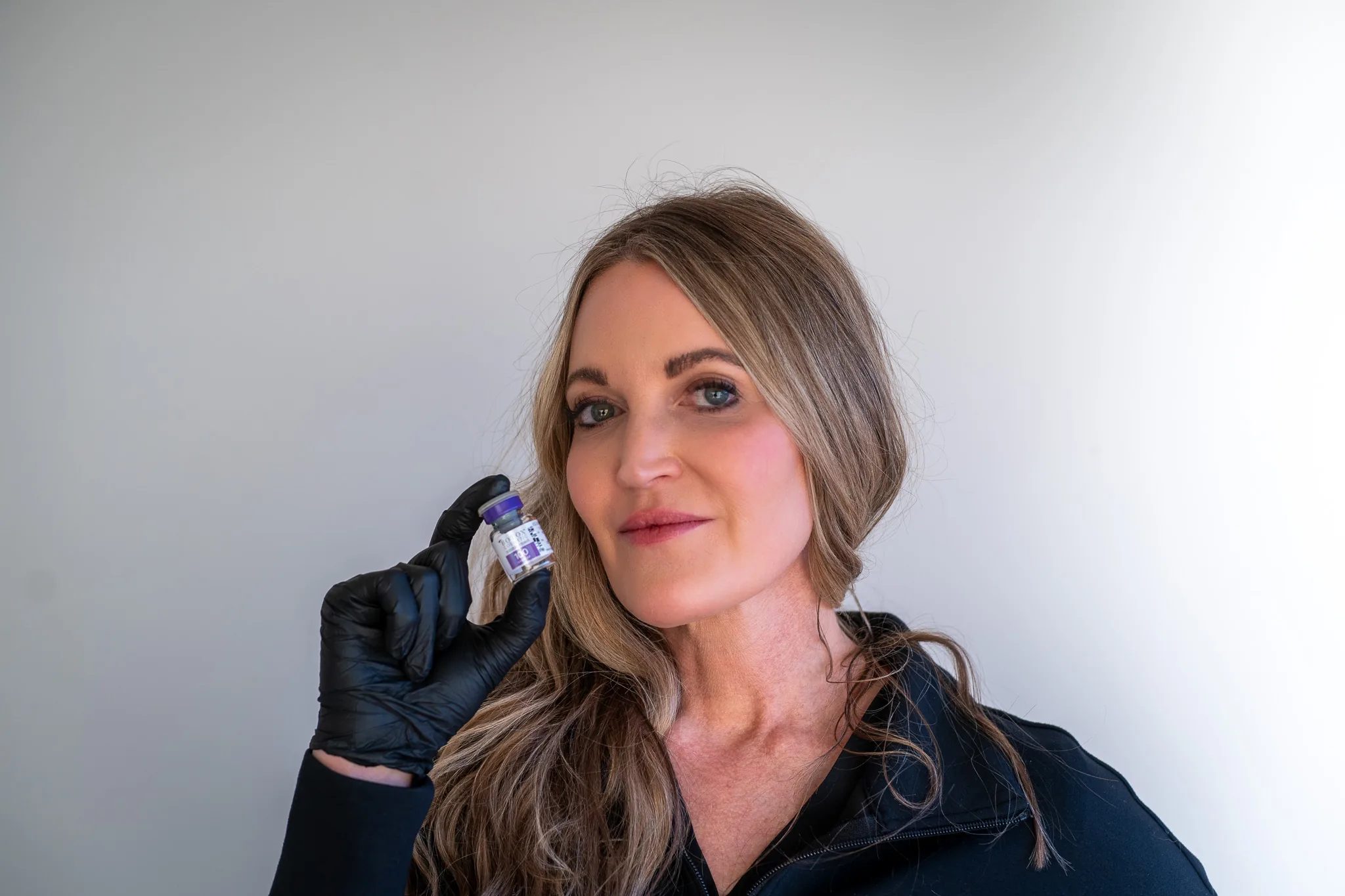 A FACE Colorado nurse in a black jacket holds a Botox Cosmetic vial up beside one cheek in a black-gloved hand.