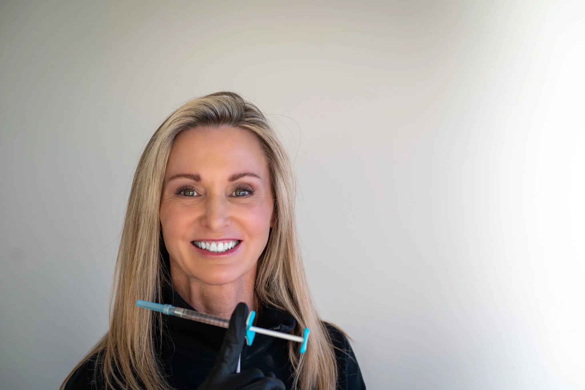 A smiling FACE Colorado nurse in a black jacket holds a syringe horizontally in a black-gloved hand against a white backdrop.
