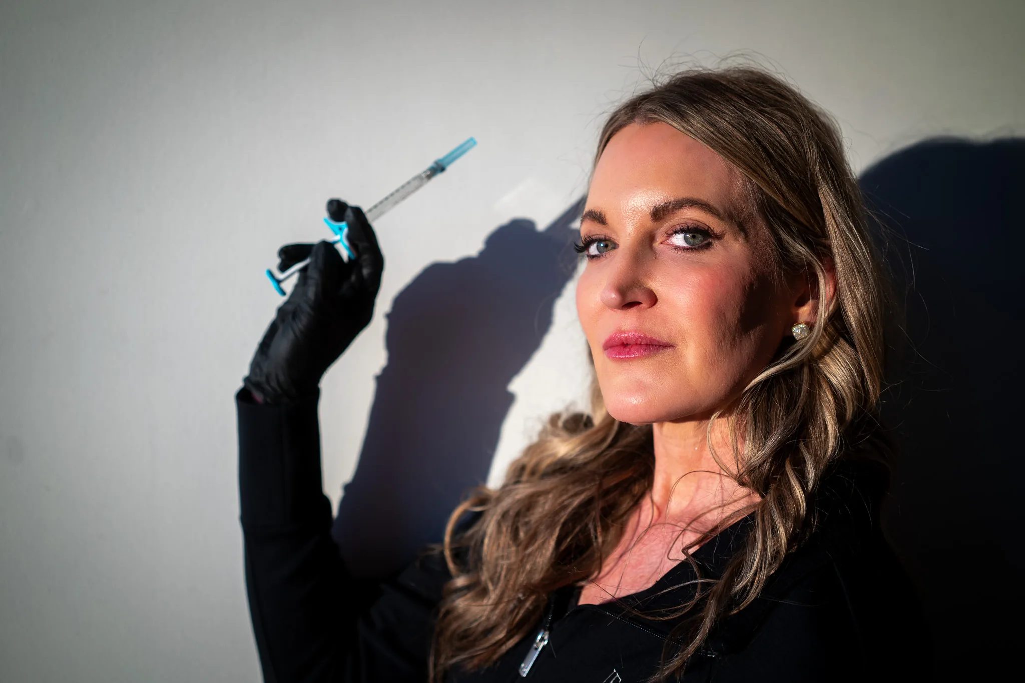 A FACE Colorado nurse in a black jacket raises a syringe in a black-gloved hand under hard light that throws a crisp shadow on the wall.
