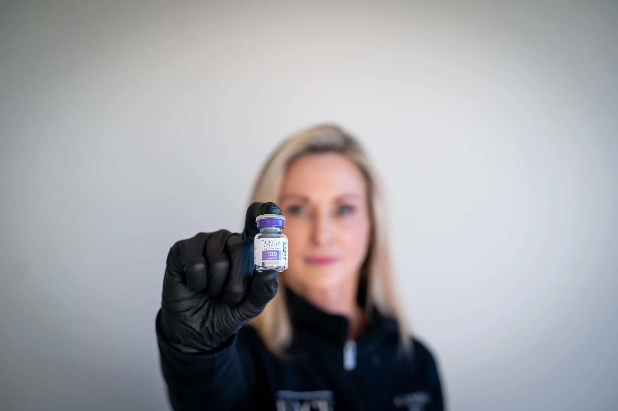 A black-gloved hand holds a Botox Cosmetic vial in sharp focus while a FACE Colorado nurse stands softly blurred behind it.
