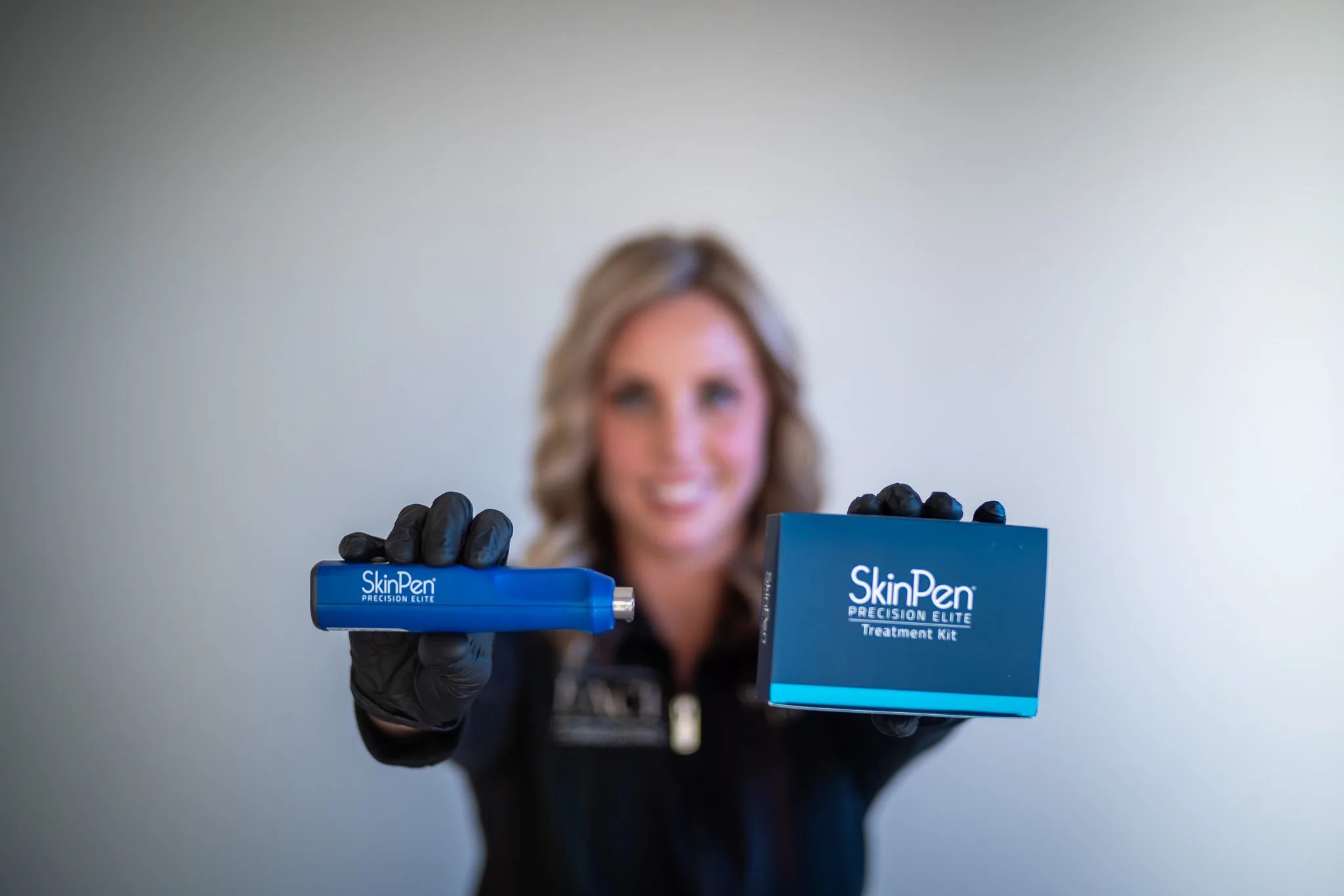 Black-gloved hands hold a SkinPen Precision Elite device and its treatment kit box in focus, with a FACE Colorado team member blurred behind.