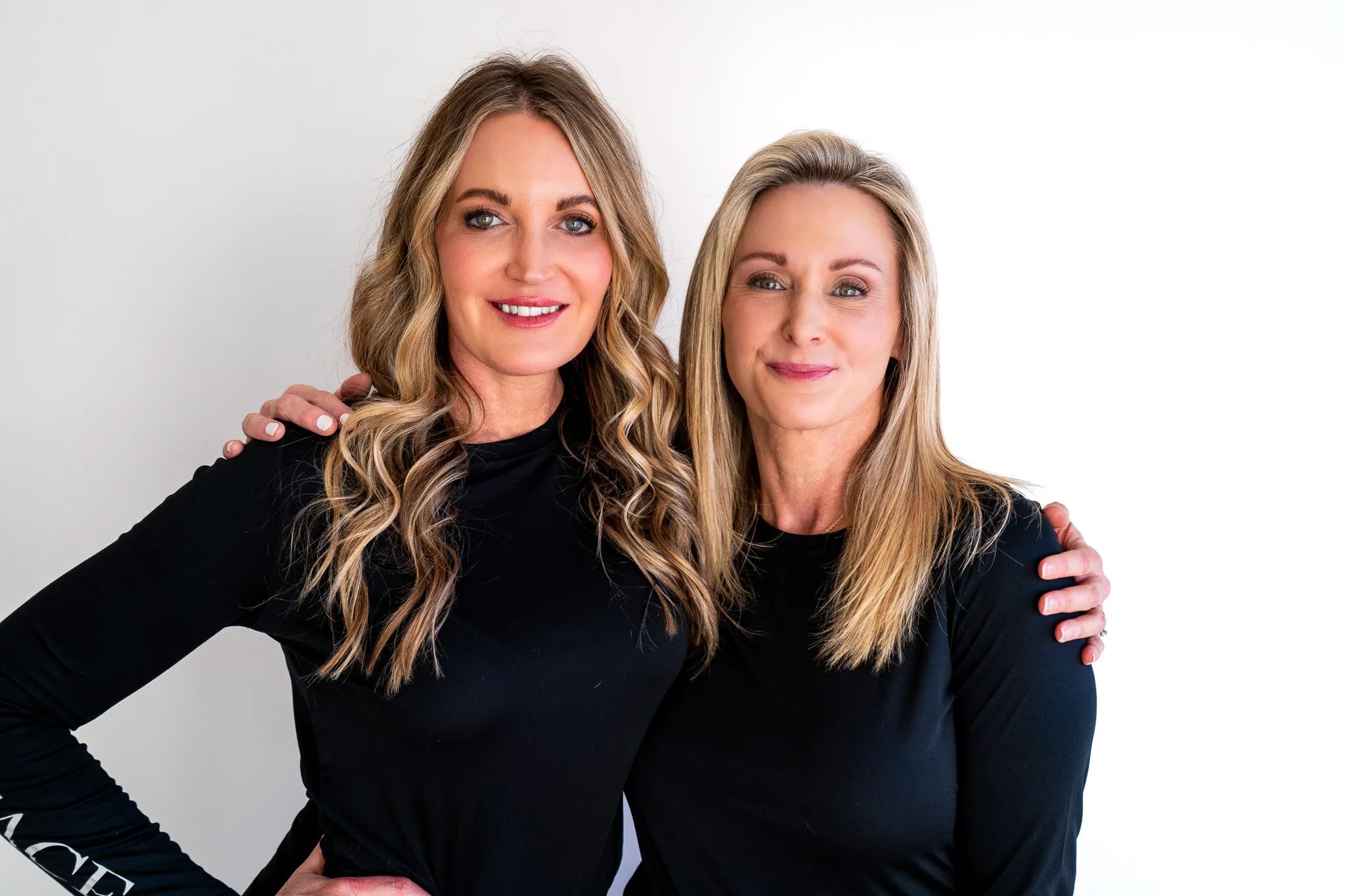 Two FACE Colorado nurses in black long-sleeve tops stand arm in arm and smile in a bright white studio.