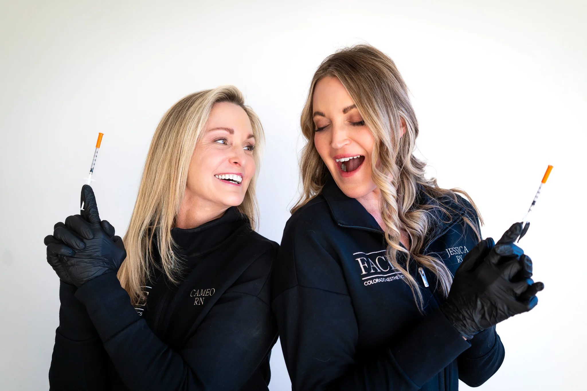 Two FACE Colorado nurses in black embroidered jackets and black gloves laugh together while each holds up a syringe.