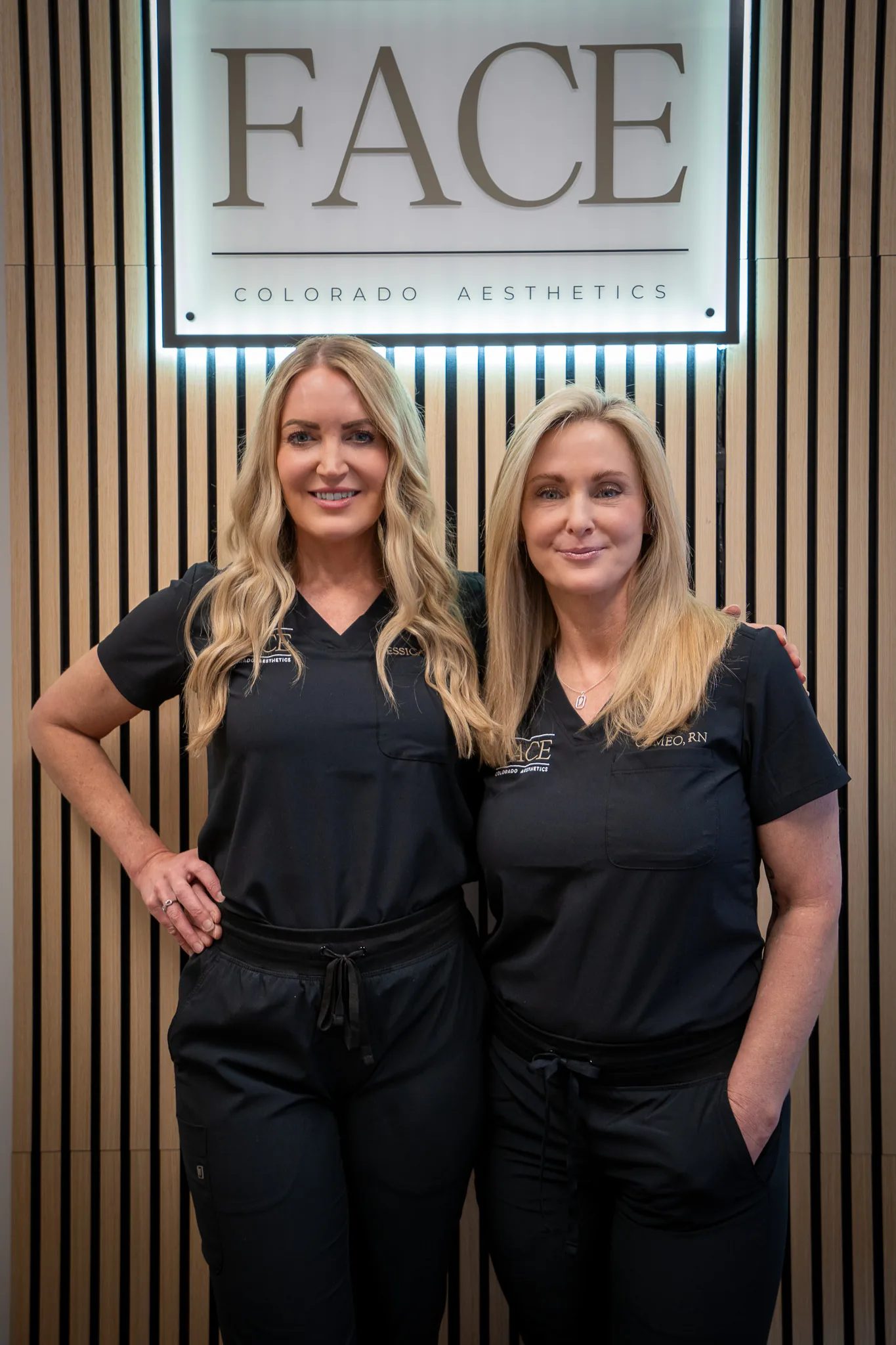 Two FACE Colorado nurses in black scrubs stand together beneath the practice's illuminated FACE Colorado Aesthetics sign.