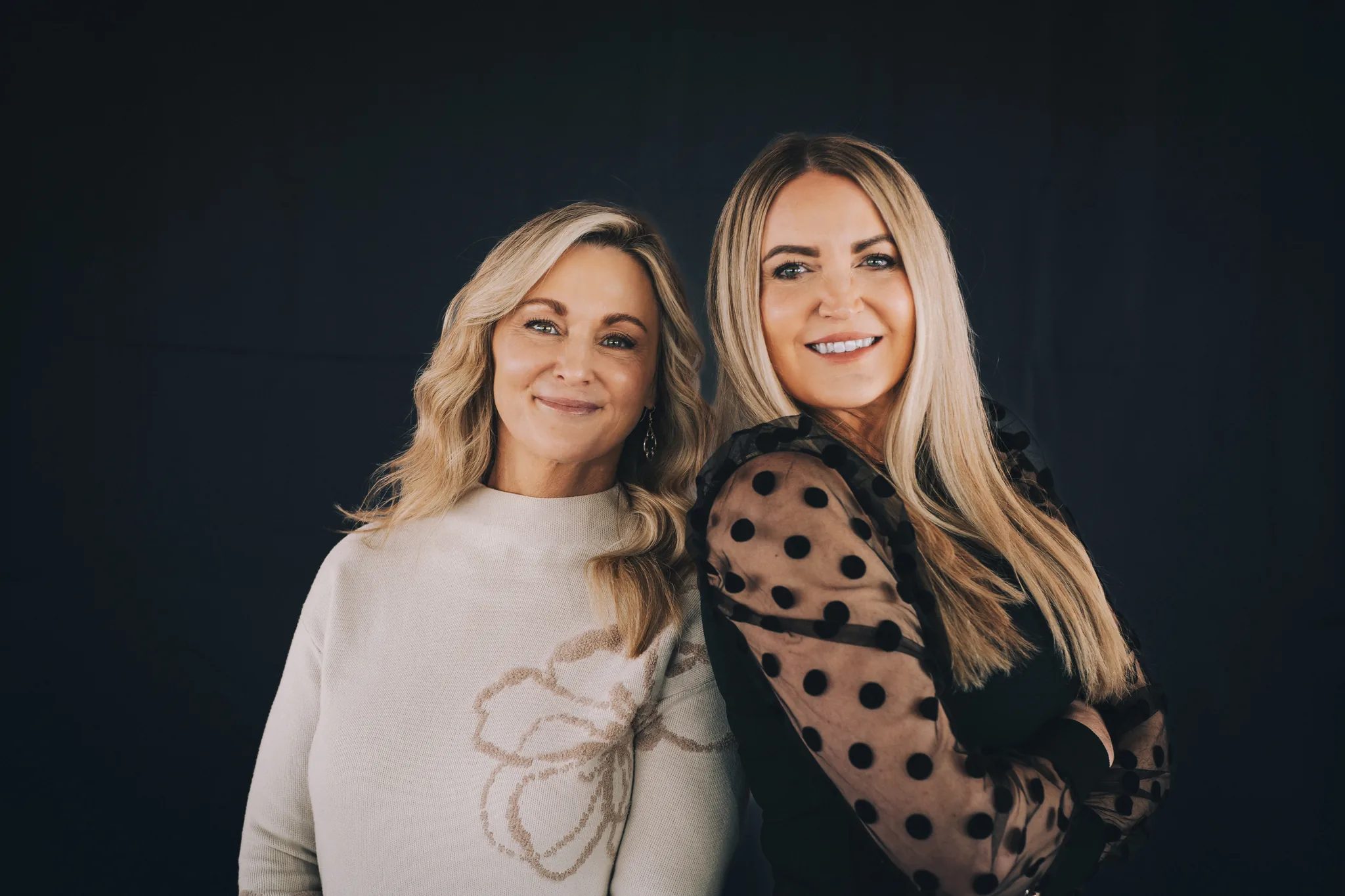 Two FACE Colorado team members pose shoulder to shoulder against a black backdrop, one in a cream floral sweater and one in a sheer polka-dot top.