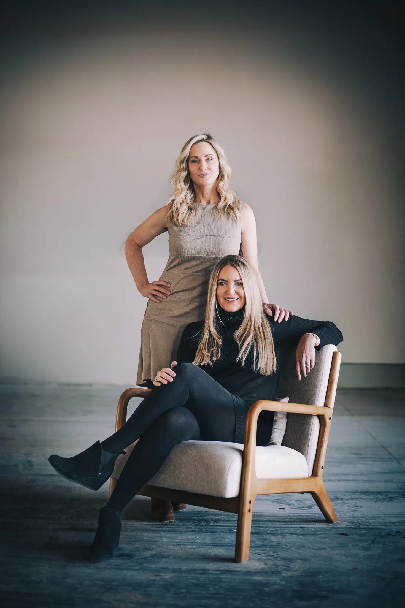 Two FACE Colorado team members pose with a wooden armchair in an open studio, one standing in a taupe dress and one seated in black.