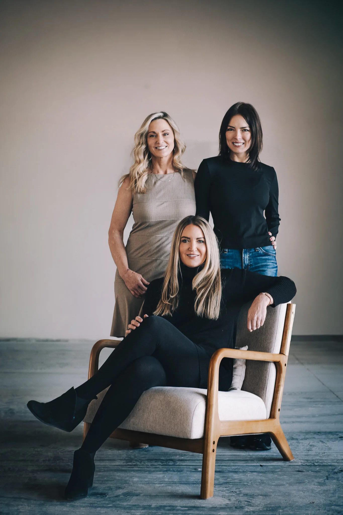 Three FACE Colorado team members pose around a wooden armchair in an open studio, two standing and one seated.