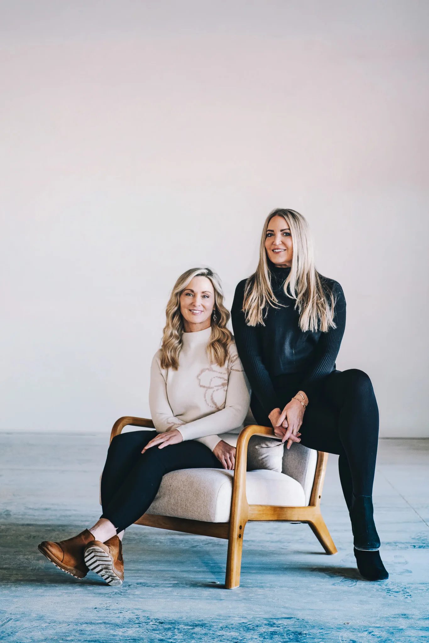 Two FACE Colorado team members share a wooden armchair, one seated in a cream sweater and one perched on the arm in black.