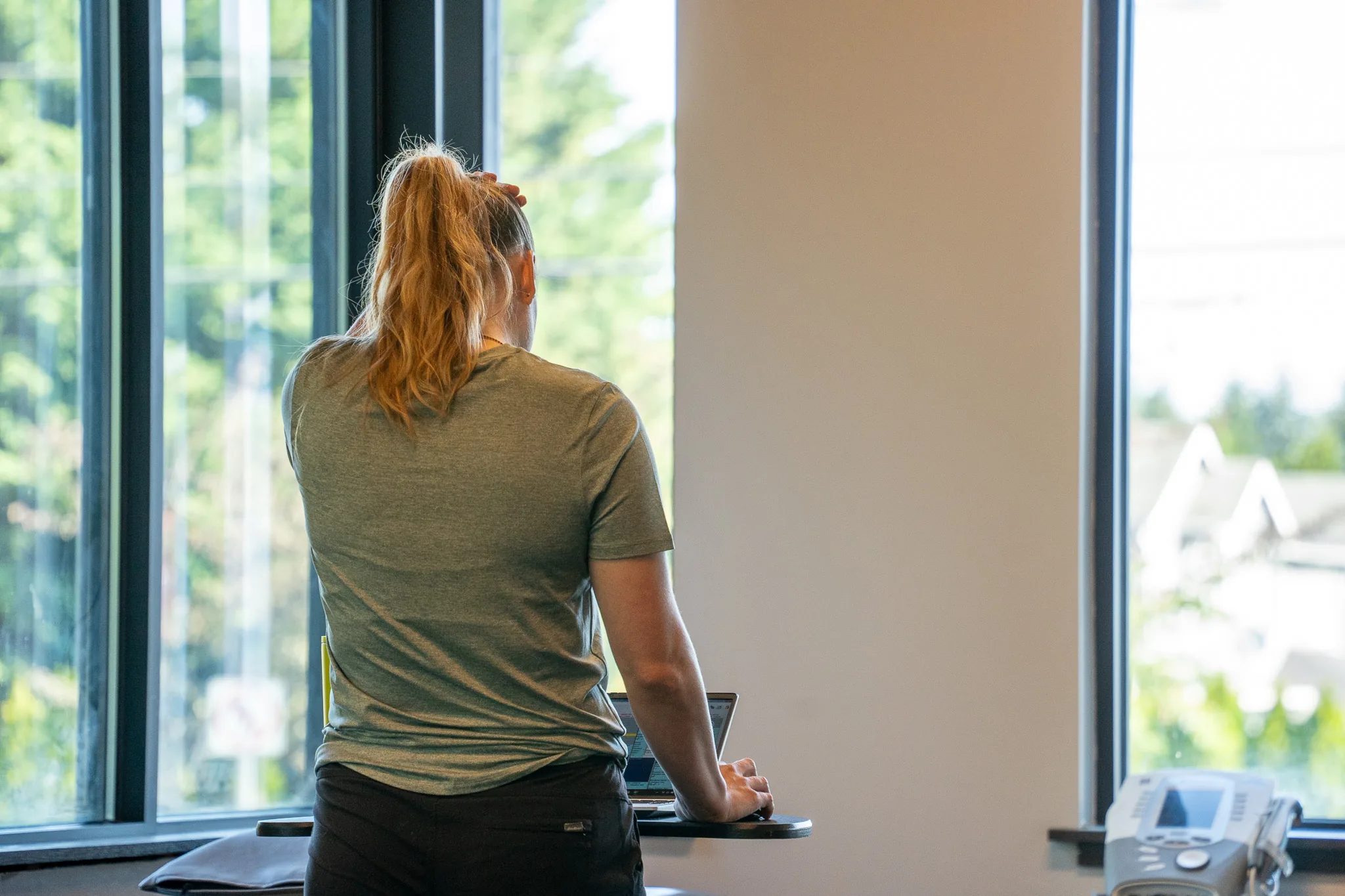 A clinician in a green T-shirt stands at a laptop beside tall windows in an IRG Physical & Hand Therapy clinic, seen from behind.