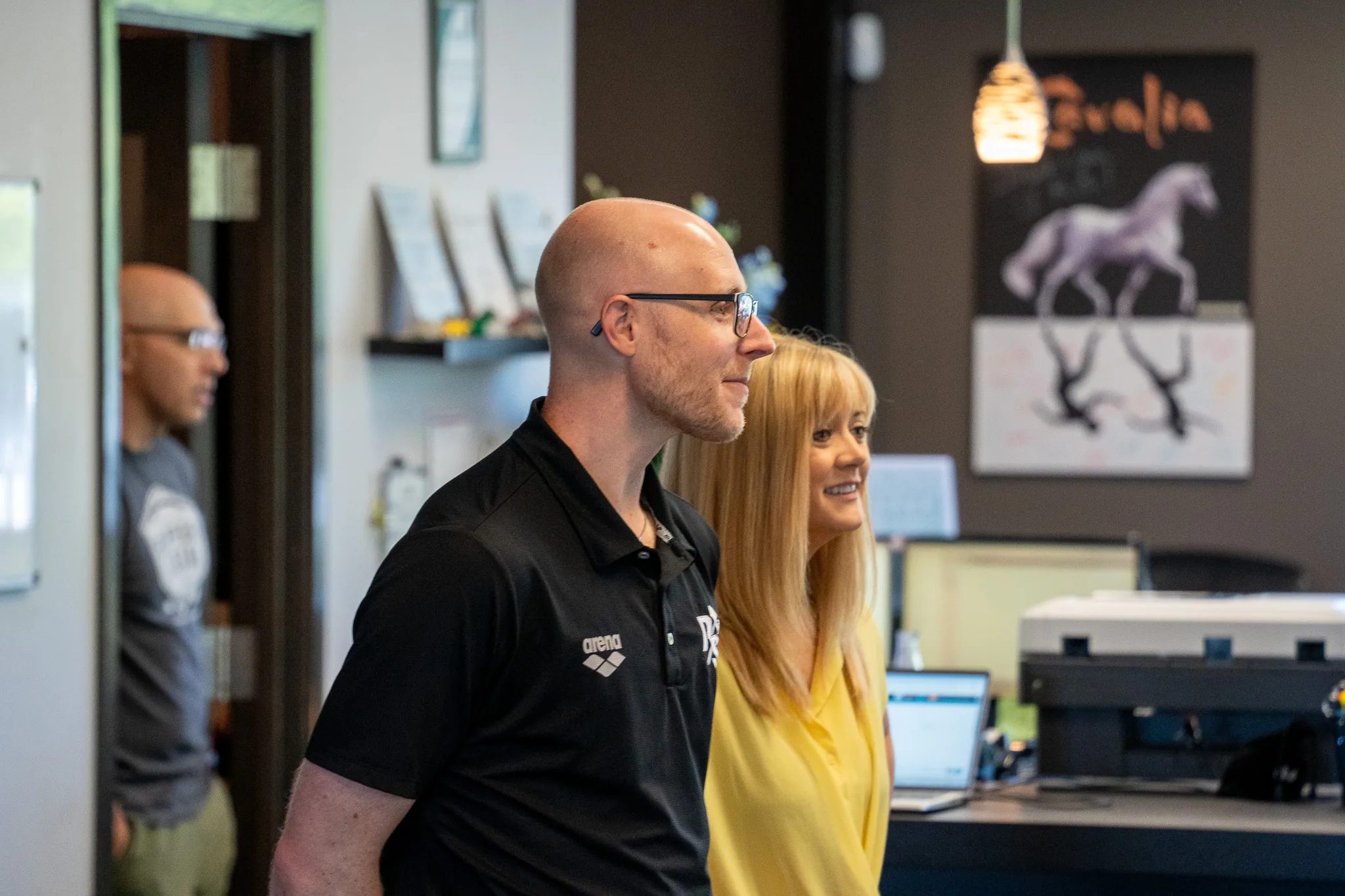 Two IRG Physical & Hand Therapy staff, a man in a black polo and a woman in yellow, smile toward a conversation near the front desk.