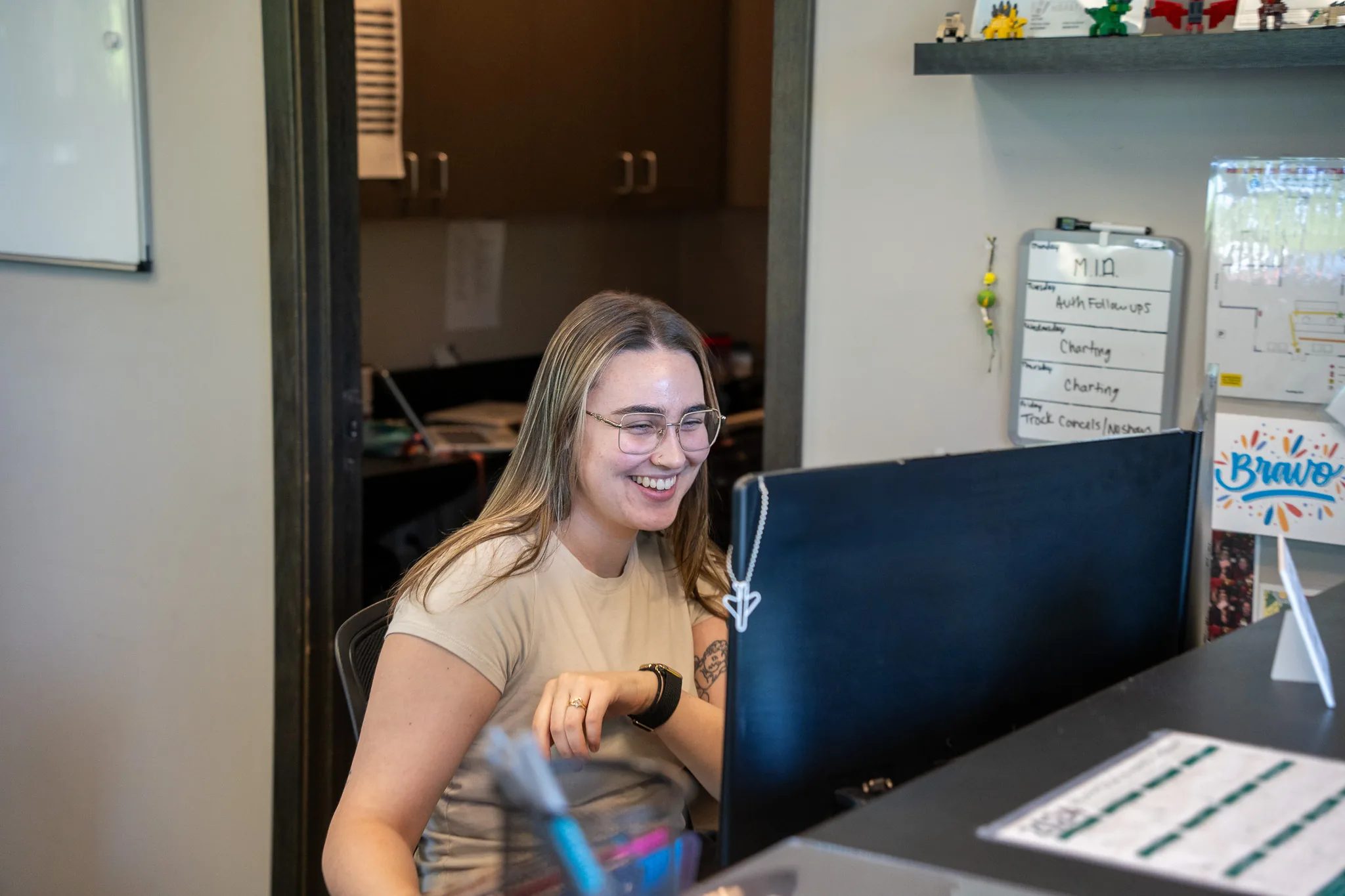 A smiling front-desk staff member in glasses works at a computer at an IRG Physical & Hand Therapy clinic.
