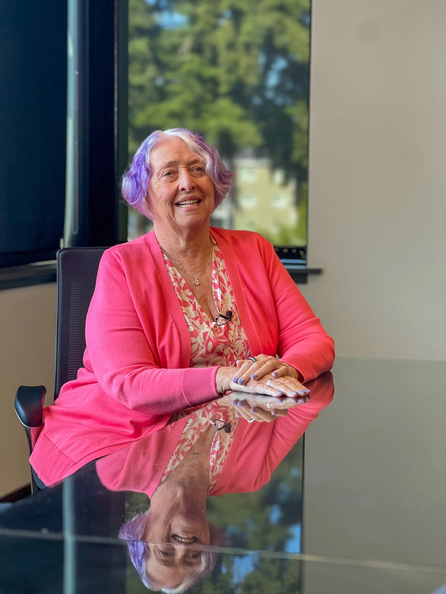A smiling woman with lavender hair and a pink cardigan sits at a glass conference table, her reflection below her.