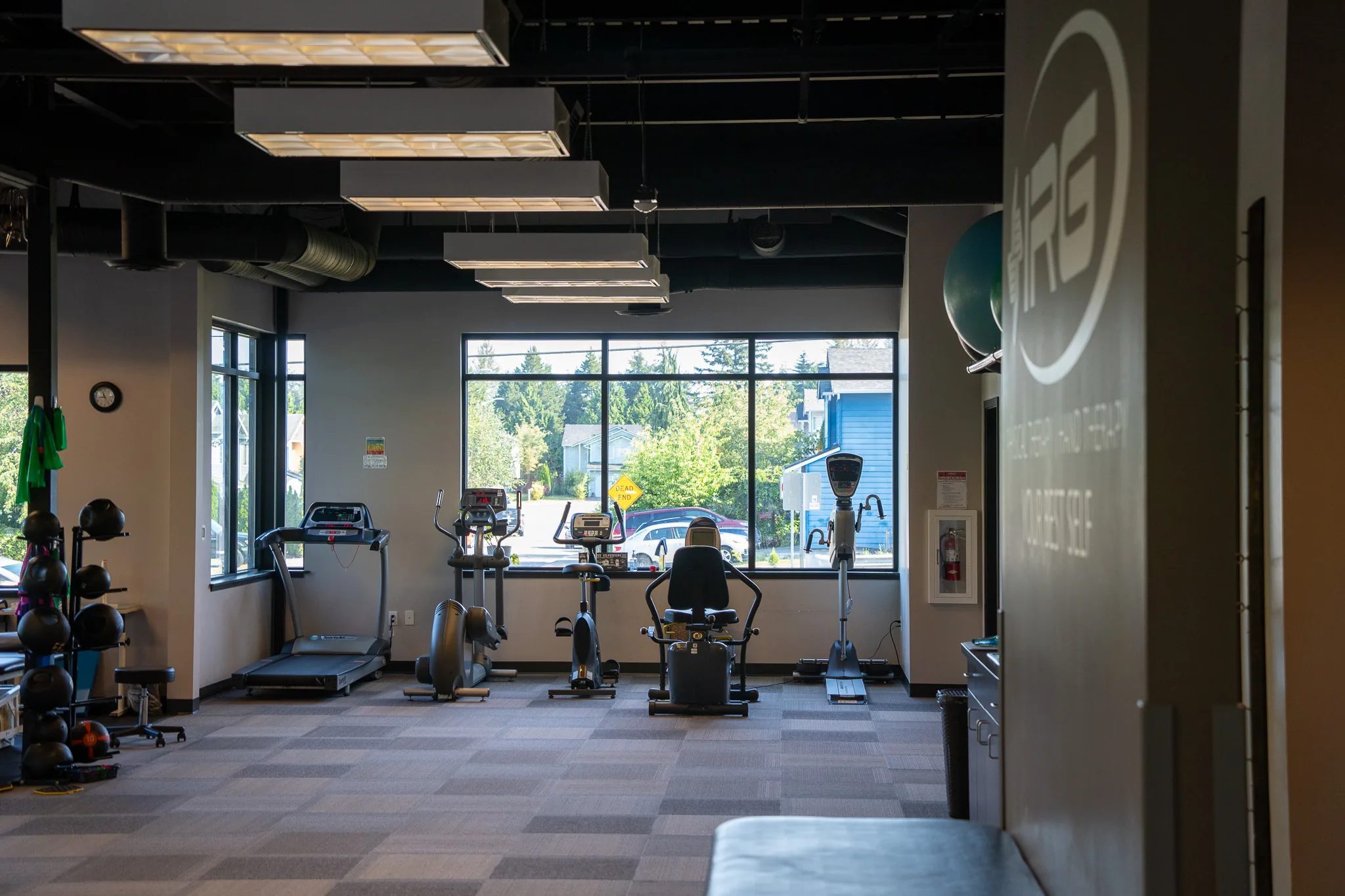 The exercise floor at an IRG Physical & Hand Therapy clinic, with a treadmill, bikes and an elliptical lined up beneath a wall of windows.