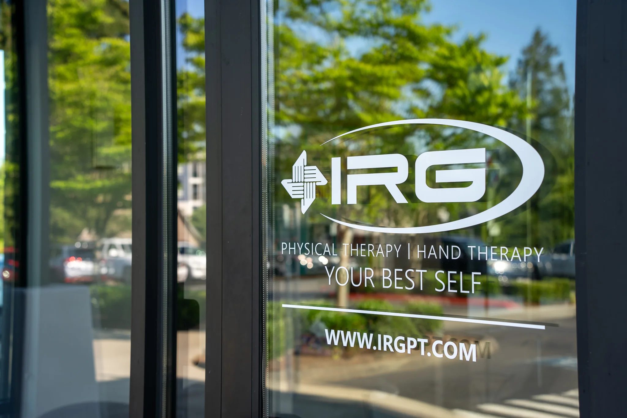 IRG Physical Therapy and Hand Therapy logo decal on a glass door reading Your Best Self and www.irgpt.com, with trees reflected in the glass.