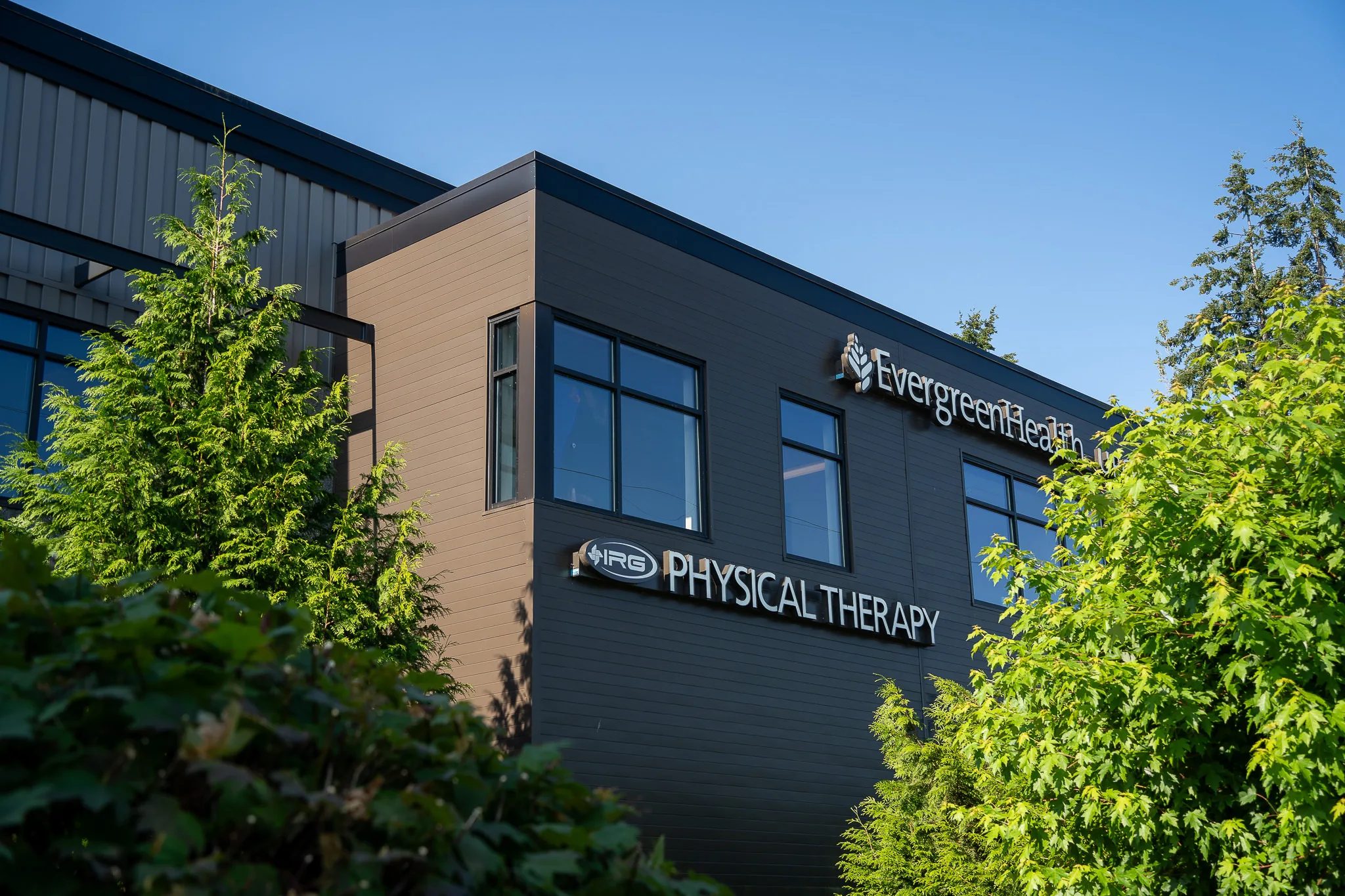 IRG Physical Therapy sign on the dark corner facade of a two-story building, framed by green trees under a clear blue sky.