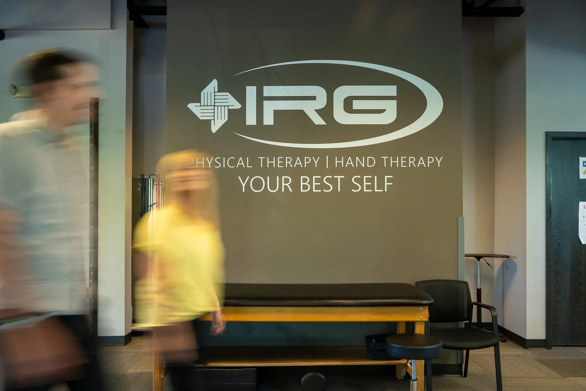 IRG Physical Therapy and Hand Therapy wall graphic reading Your Best Self, with two motion-blurred people walking past a treatment table.
