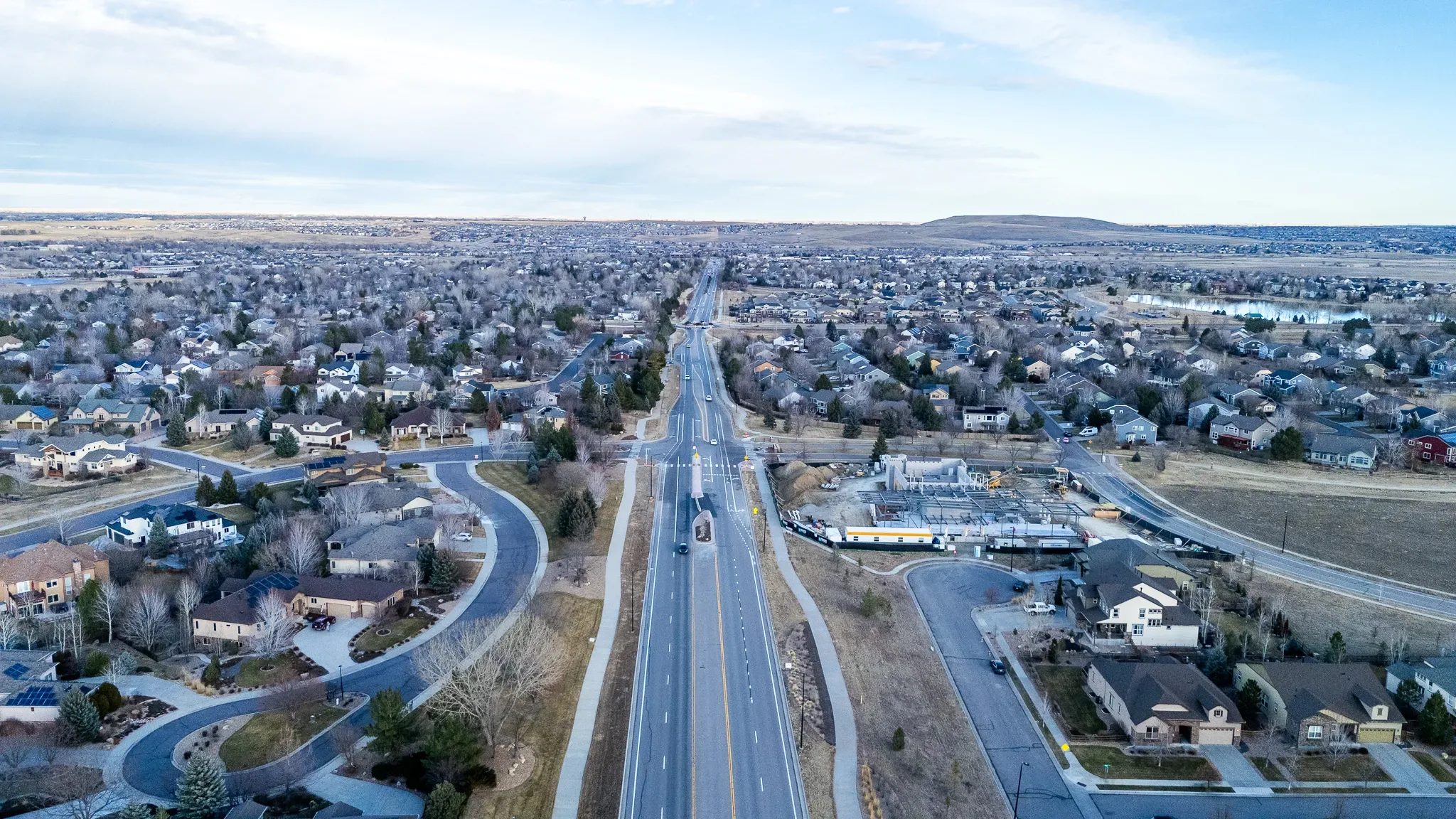 Aerial view down a divided road through a Denver-area subdivision, with a steel-framed building under construction at right.