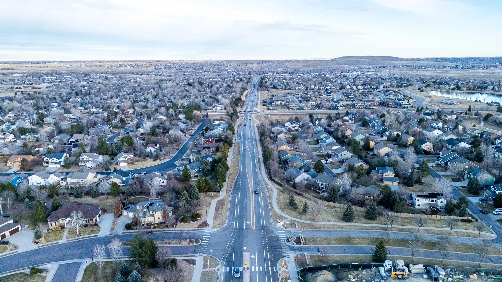 Wide drone view along a divided road through a Denver-area subdivision toward a distant mesa, with a construction staging yard at lower right.