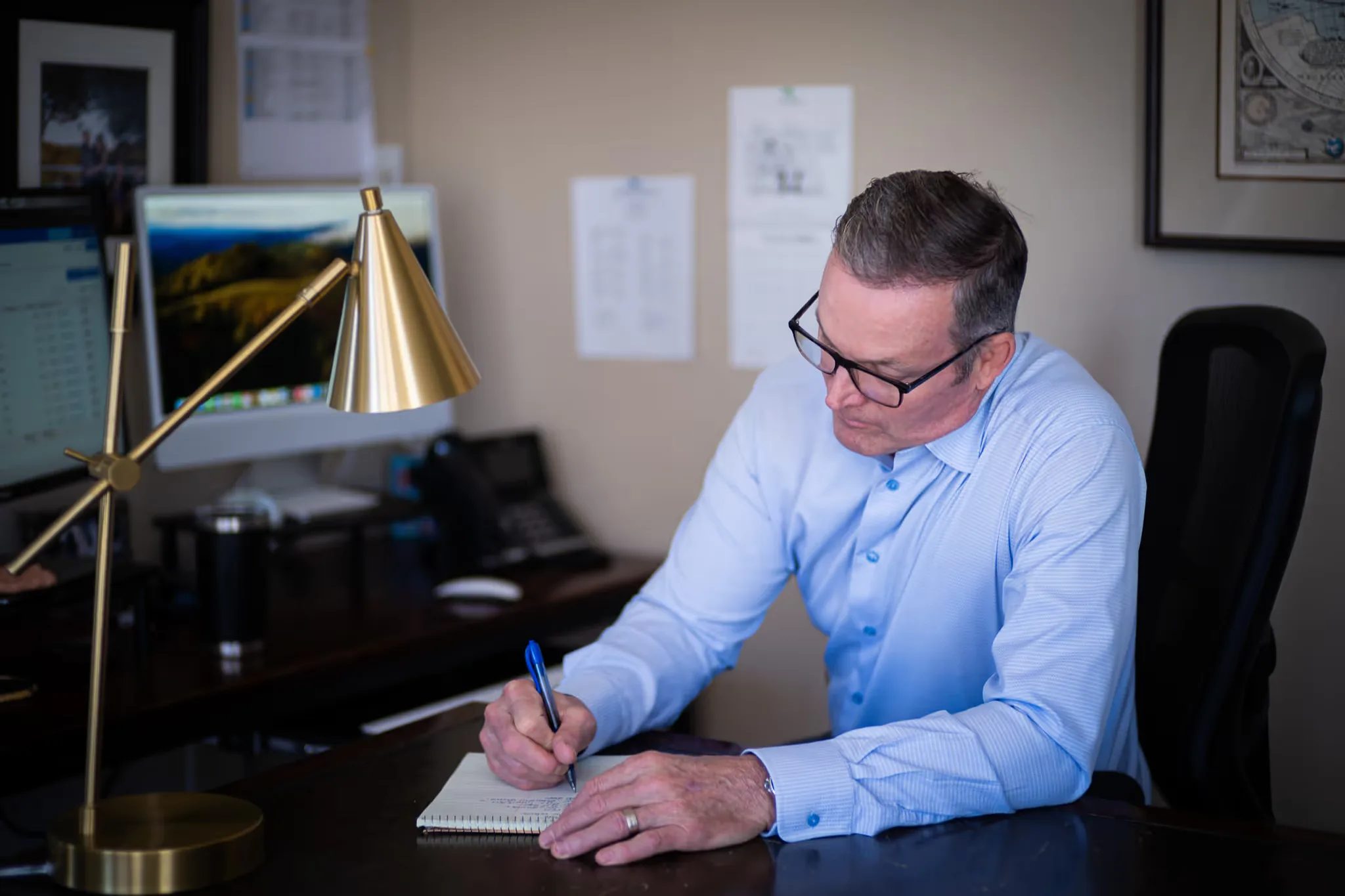 A staff member writes on a notepad at a dark wood desk under a brass cone lamp at Nelson Law Firm.