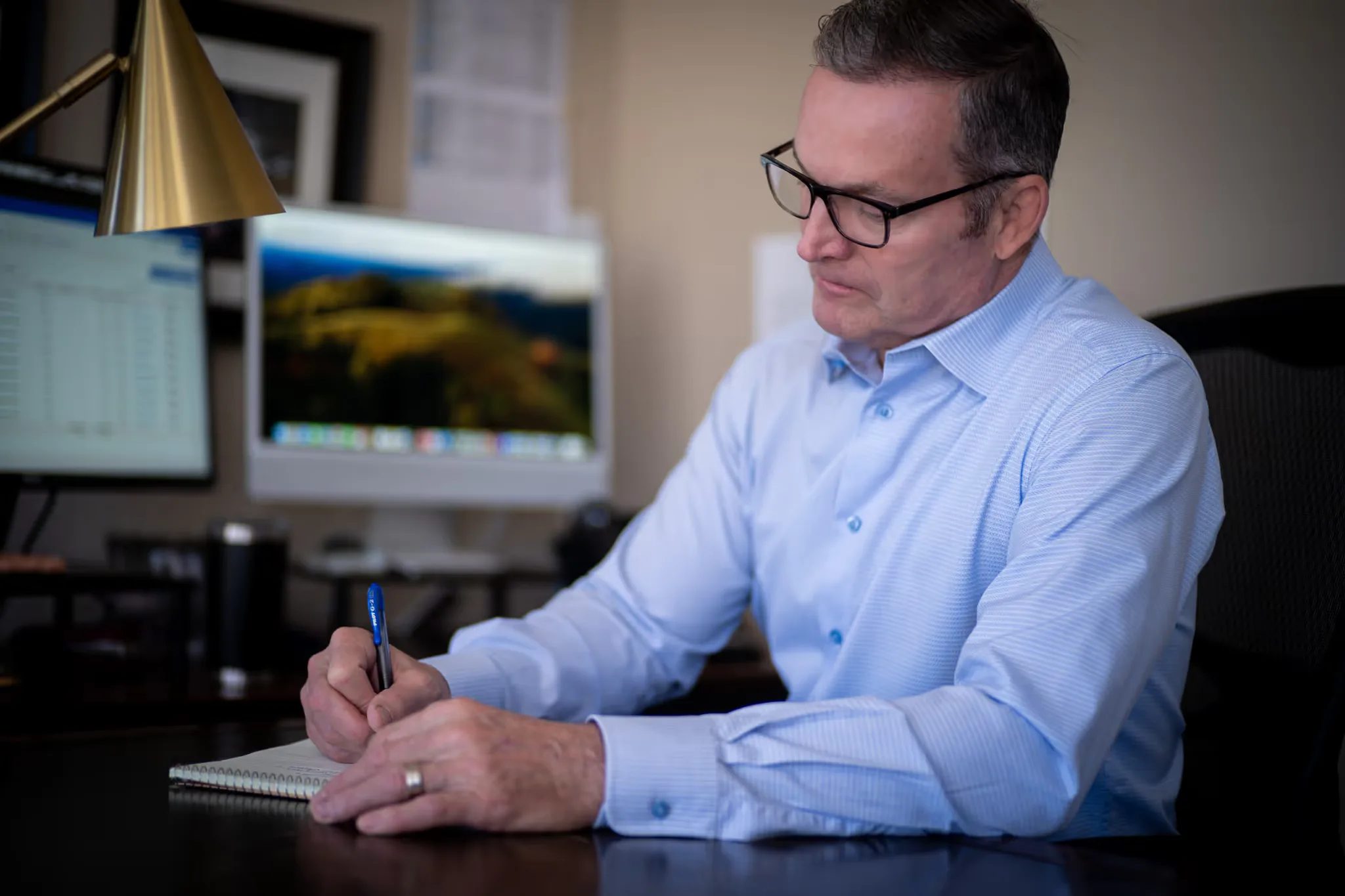 A staff member with glasses writes on a notepad at a desk with a brass cone lamp and monitors at Nelson Law Firm.