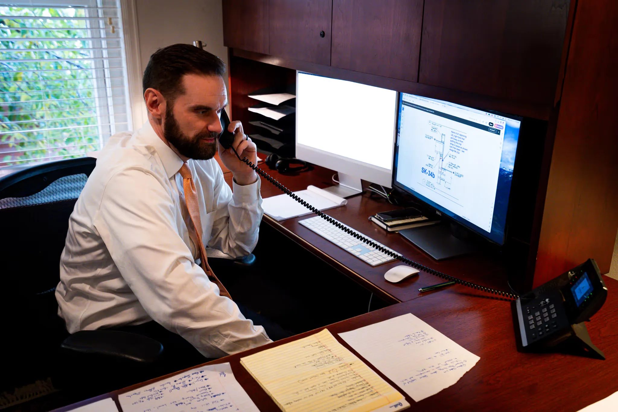 A staff member takes a phone call at a desk with dual monitors, a technical drawing on screen, at Nelson Law Firm.