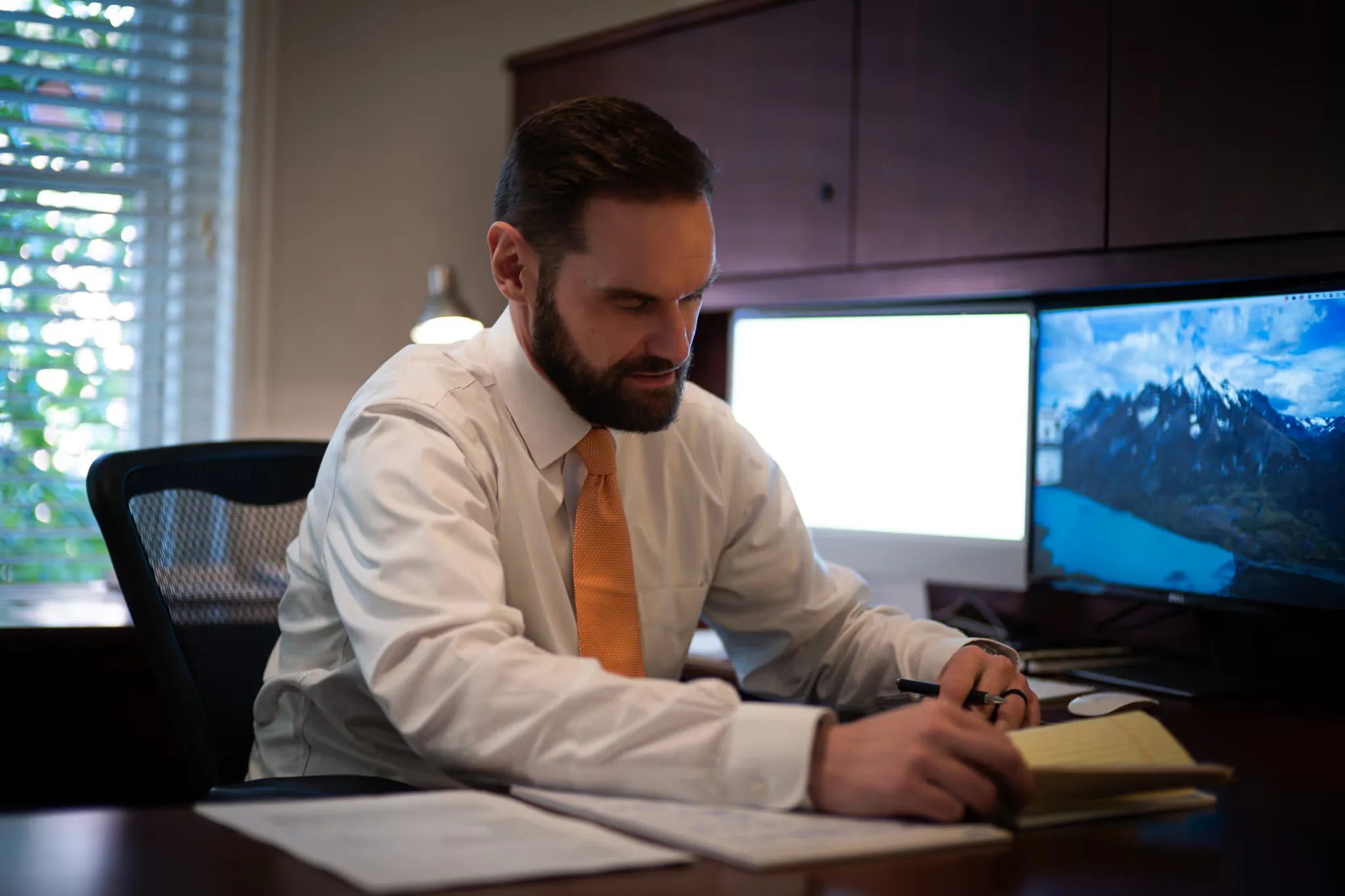 A staff member in a white shirt and orange tie writes on a yellow legal pad at a desk with dual monitors at Nelson Law Firm.