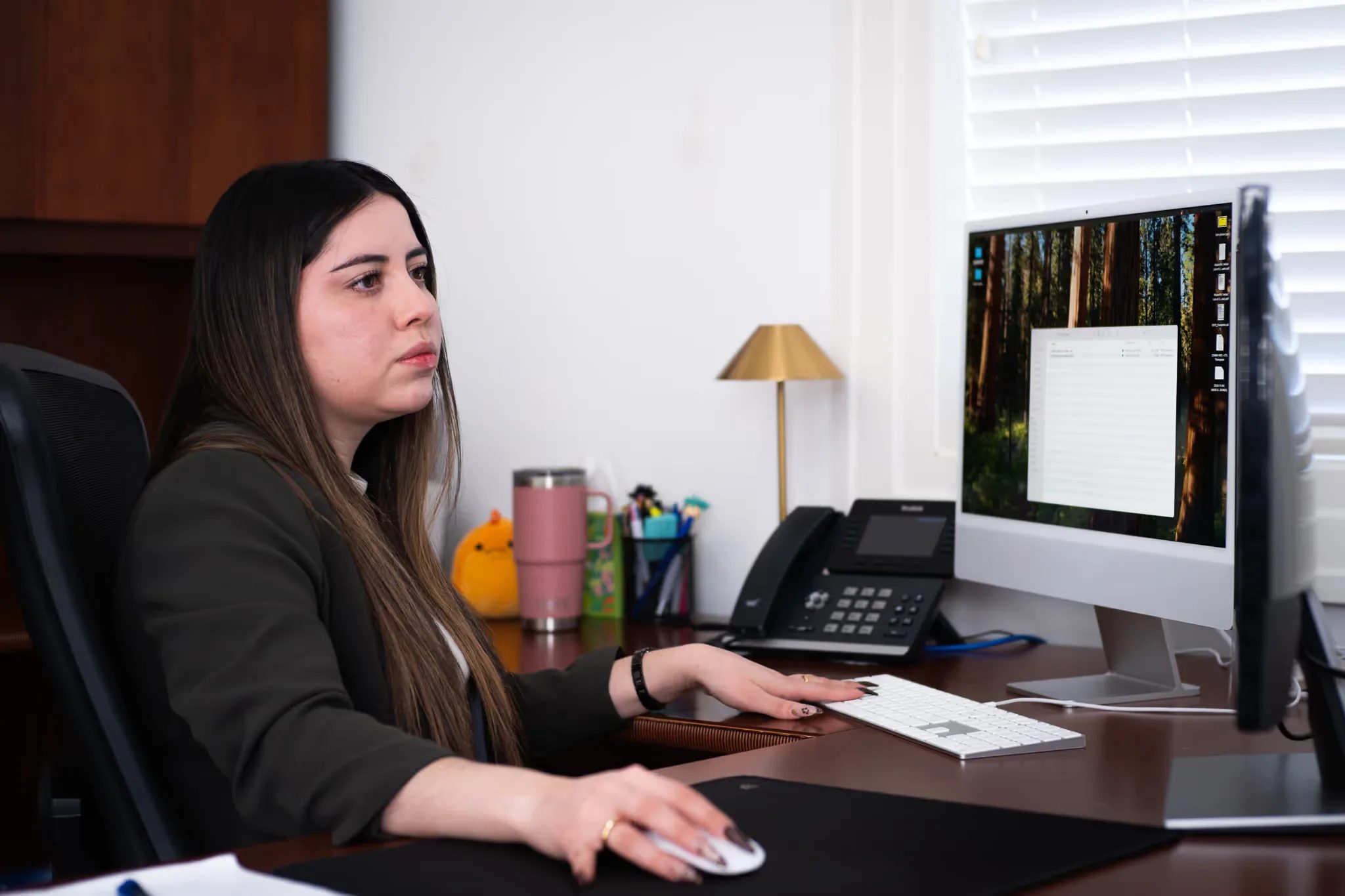 Mariana Gonzalez, Paralegal at Nelson Law Firm, looks at an iMac screen beside a desk phone and a brass lamp.