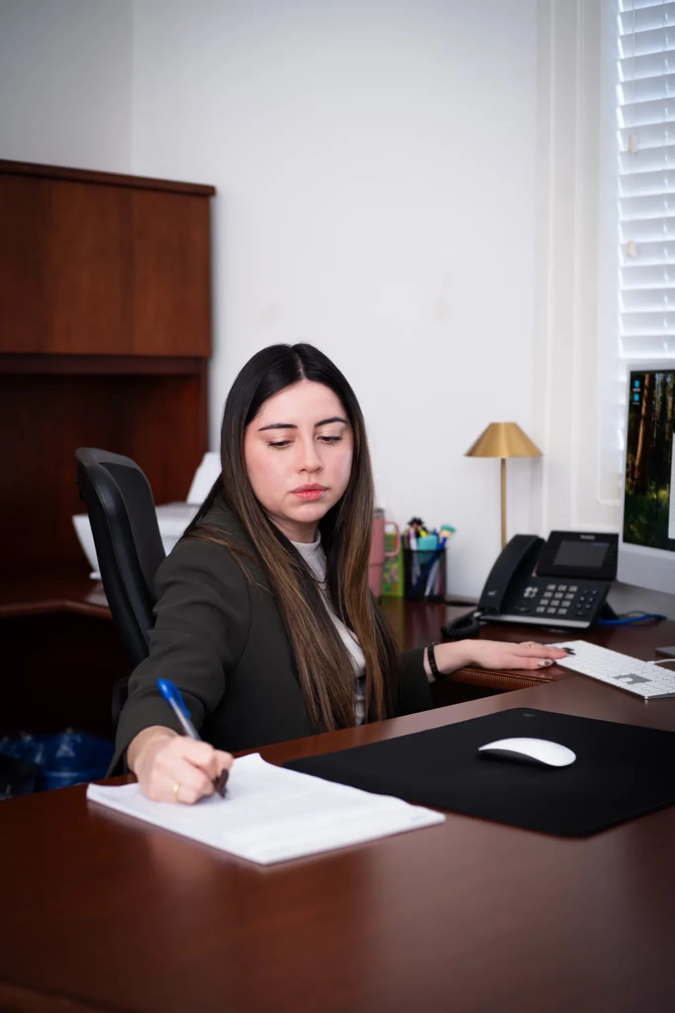 Mariana Gonzalez, Paralegal, writes on a document at a desk with an iMac and desk phone at Nelson Law Firm.