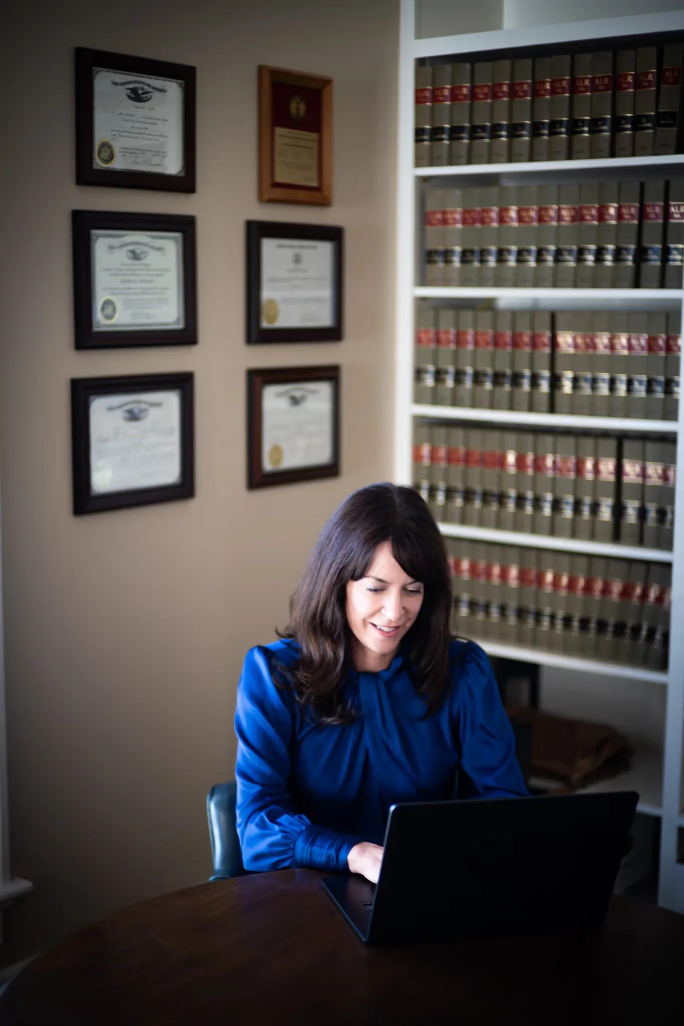 Colleen S. Nelson at a laptop beside shelves of law books and framed diplomas in the Nelson Law Firm library