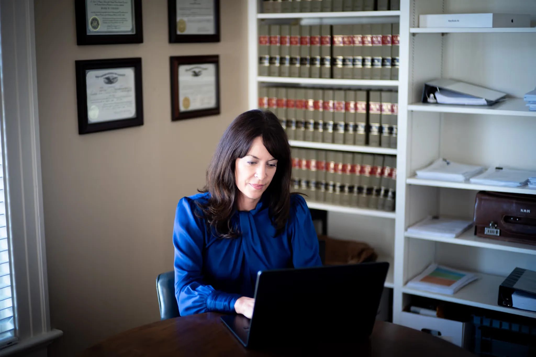 Wide view of Colleen S. Nelson working on a laptop in the Nelson Law Firm library, with binders, law books and framed certificates