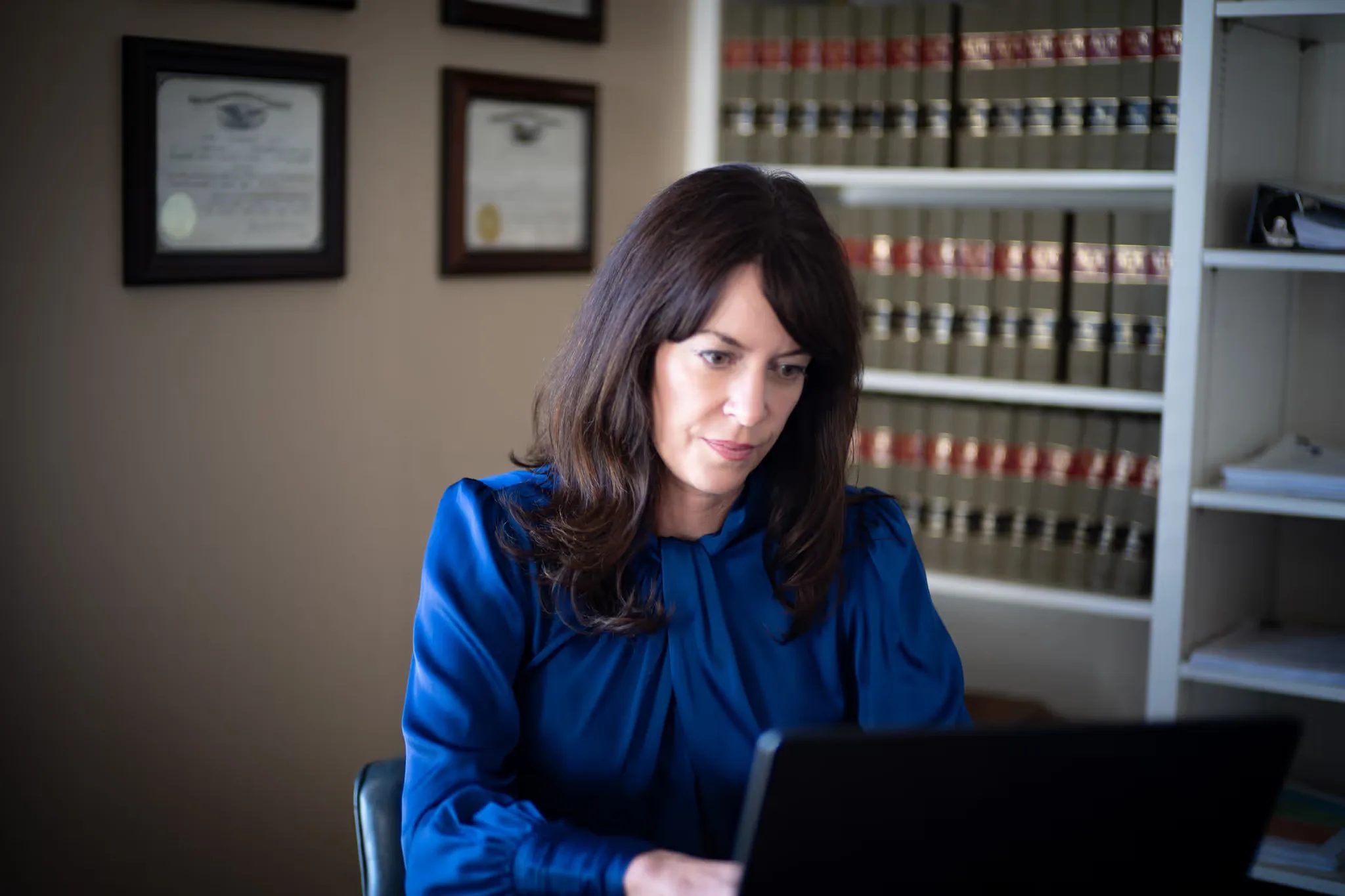 Close view of Colleen S. Nelson concentrating on a laptop screen, with framed certificates and law books behind, at Nelson Law Firm