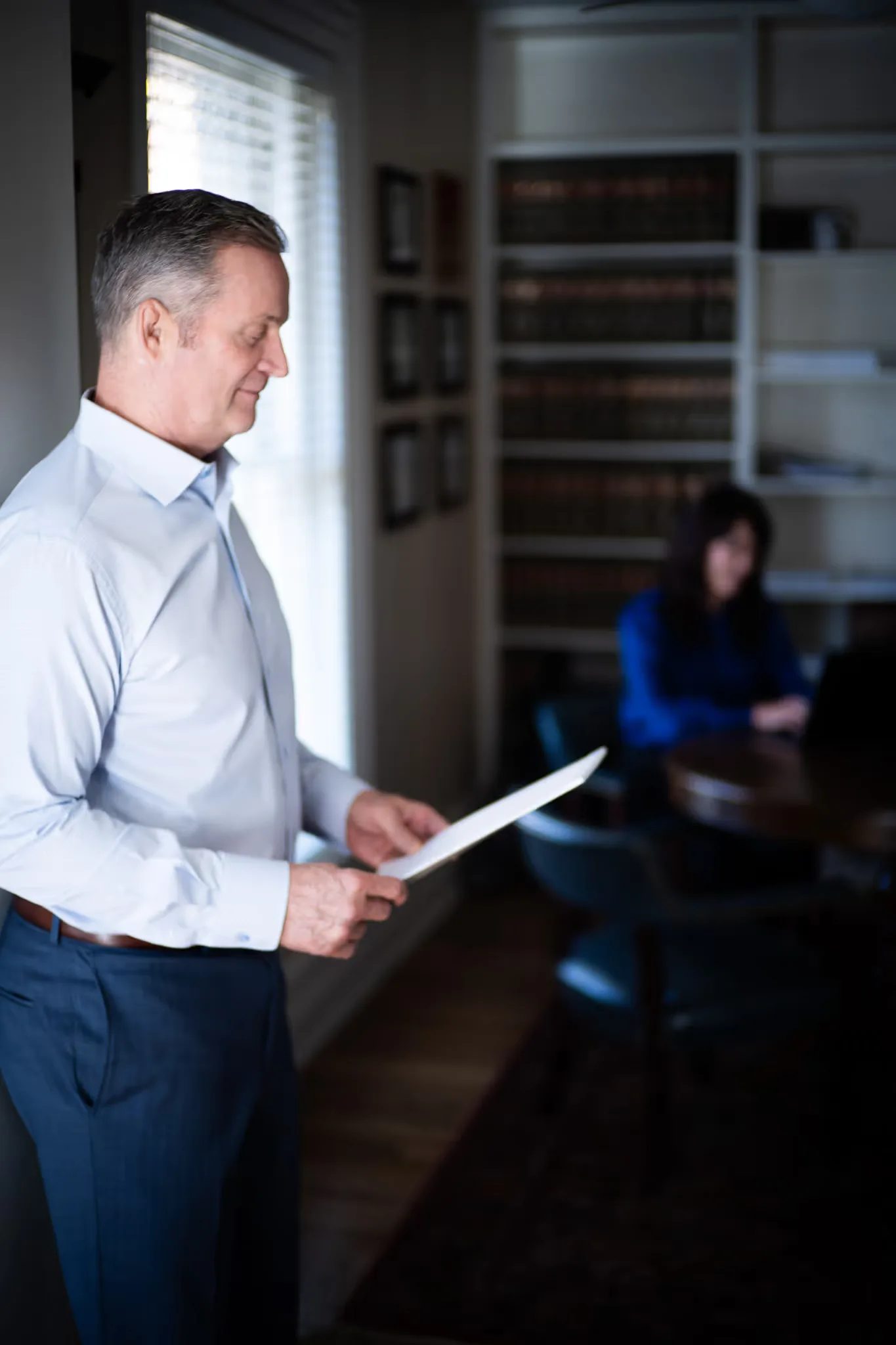 A staff member in a light blue shirt reads a document by a window while a colleague works at a laptop in the Nelson Law Firm library.