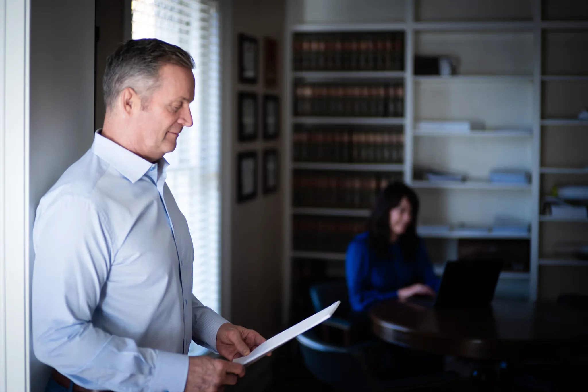 A staff member reads a document beside a window in the Nelson Law Firm library while a colleague works at a laptop behind.