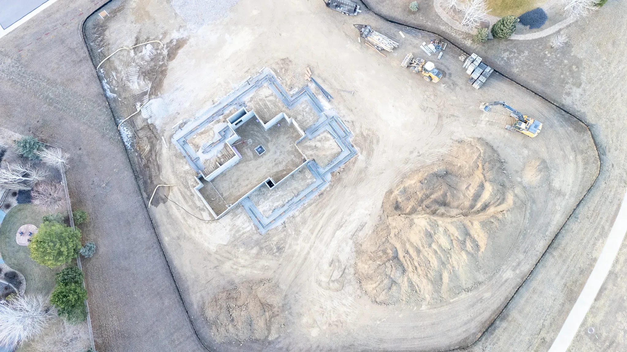 Top-down drone view of a new home's poured concrete foundation with an excavator and loader on a graded lot.