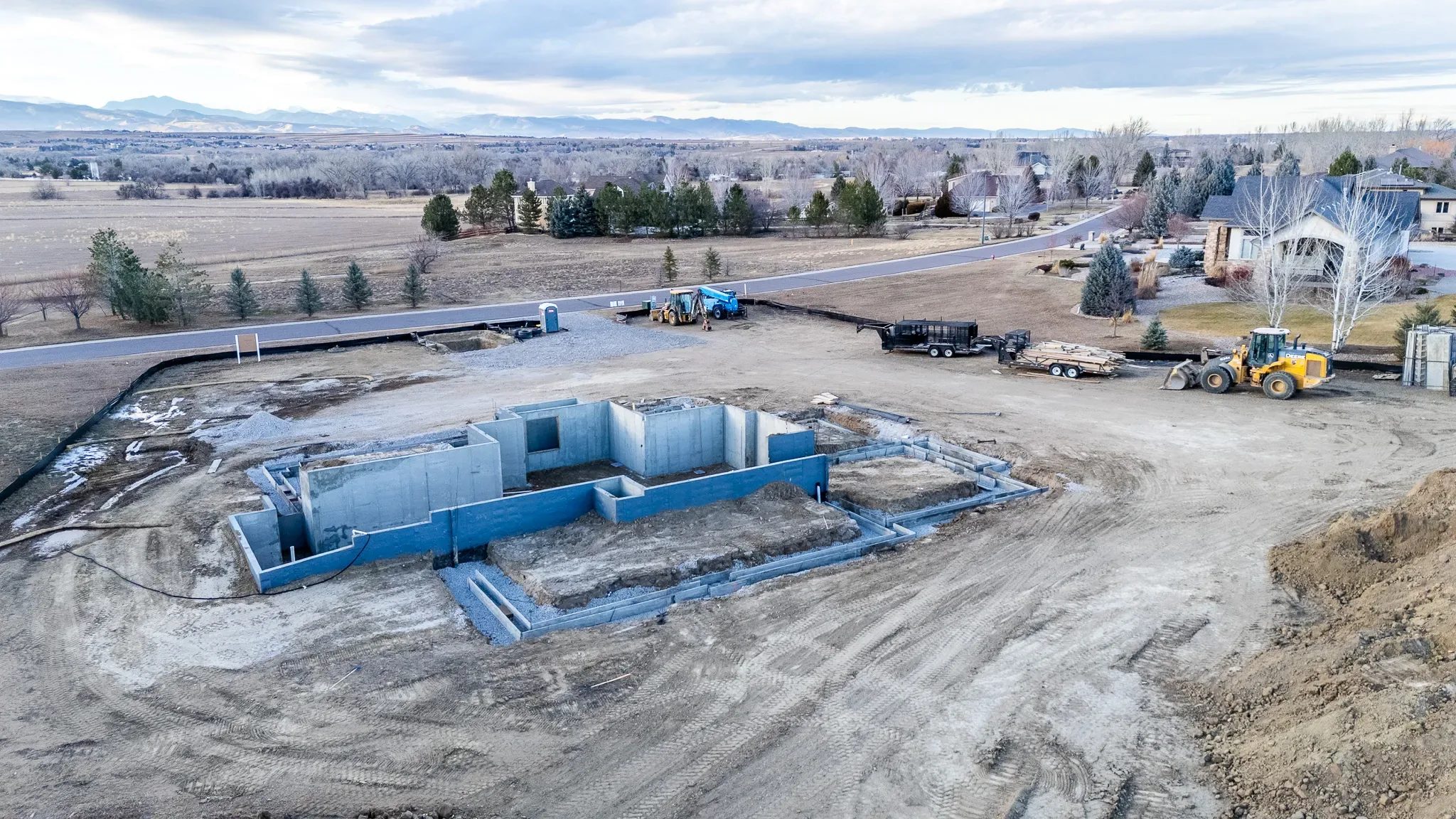 Aerial view of a residential foundation under construction with concrete walls, a loader, a trailer of lumber and mountains beyond.