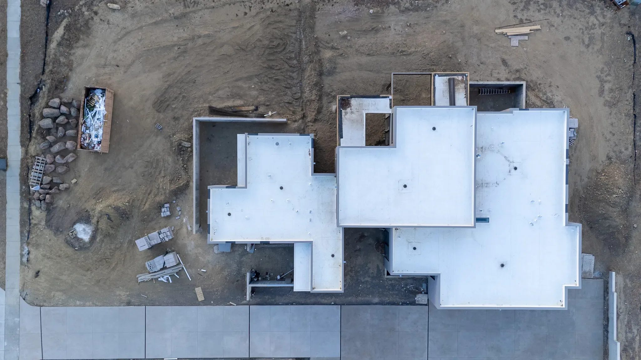 Top-down drone view of a modern home under construction with white flat roofs, stacked materials and a dumpster.