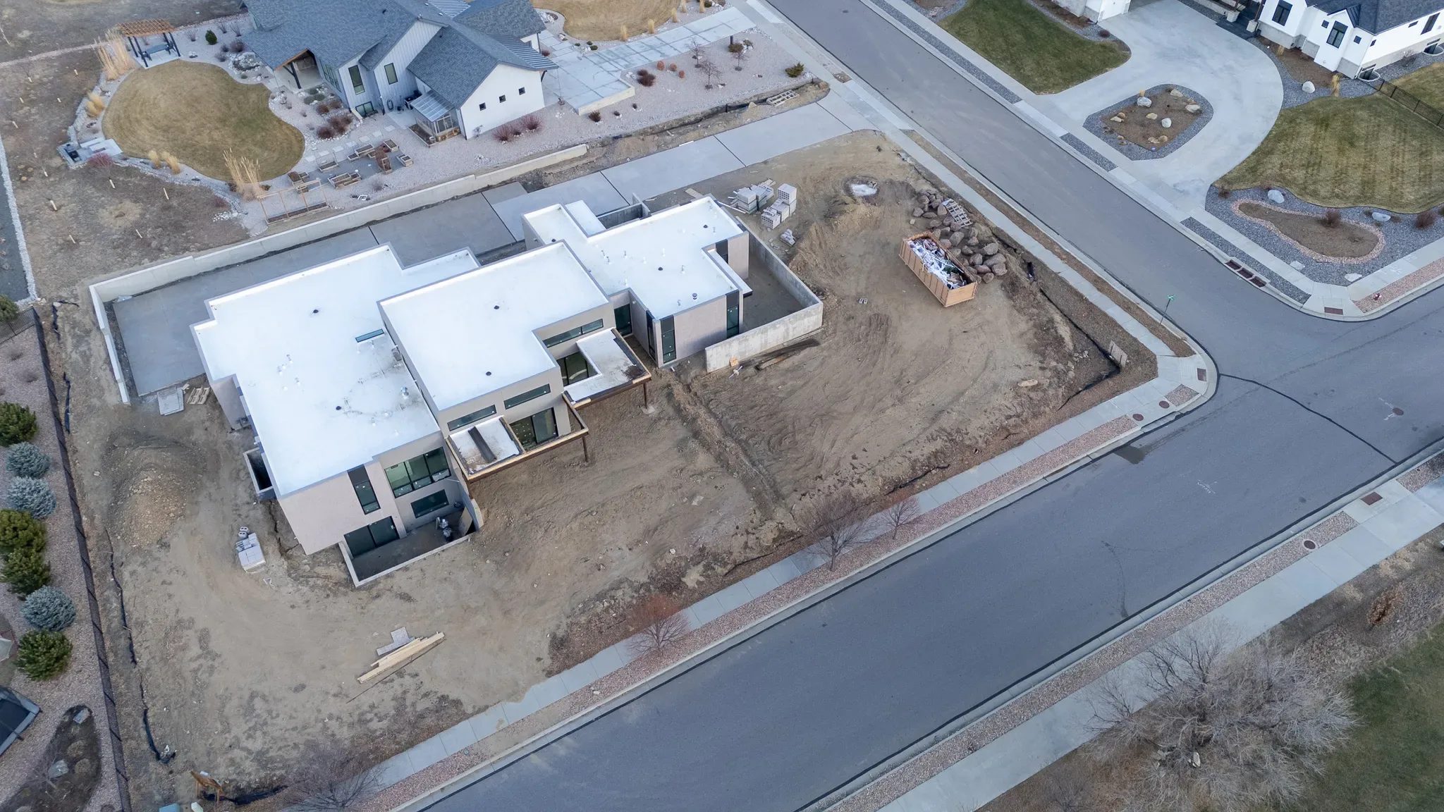 High-angle aerial of a contemporary home under construction on a corner lot, with sidewalks, street and a neighboring farmhouse-style home.