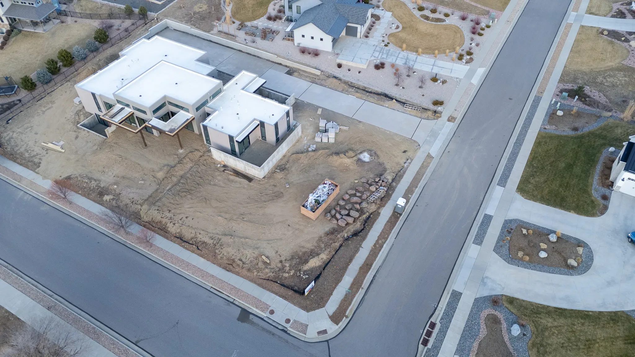 Aerial view of a modern home under construction on a corner lot, with a dumpster, boulders and dirt yard, neighbors beyond.