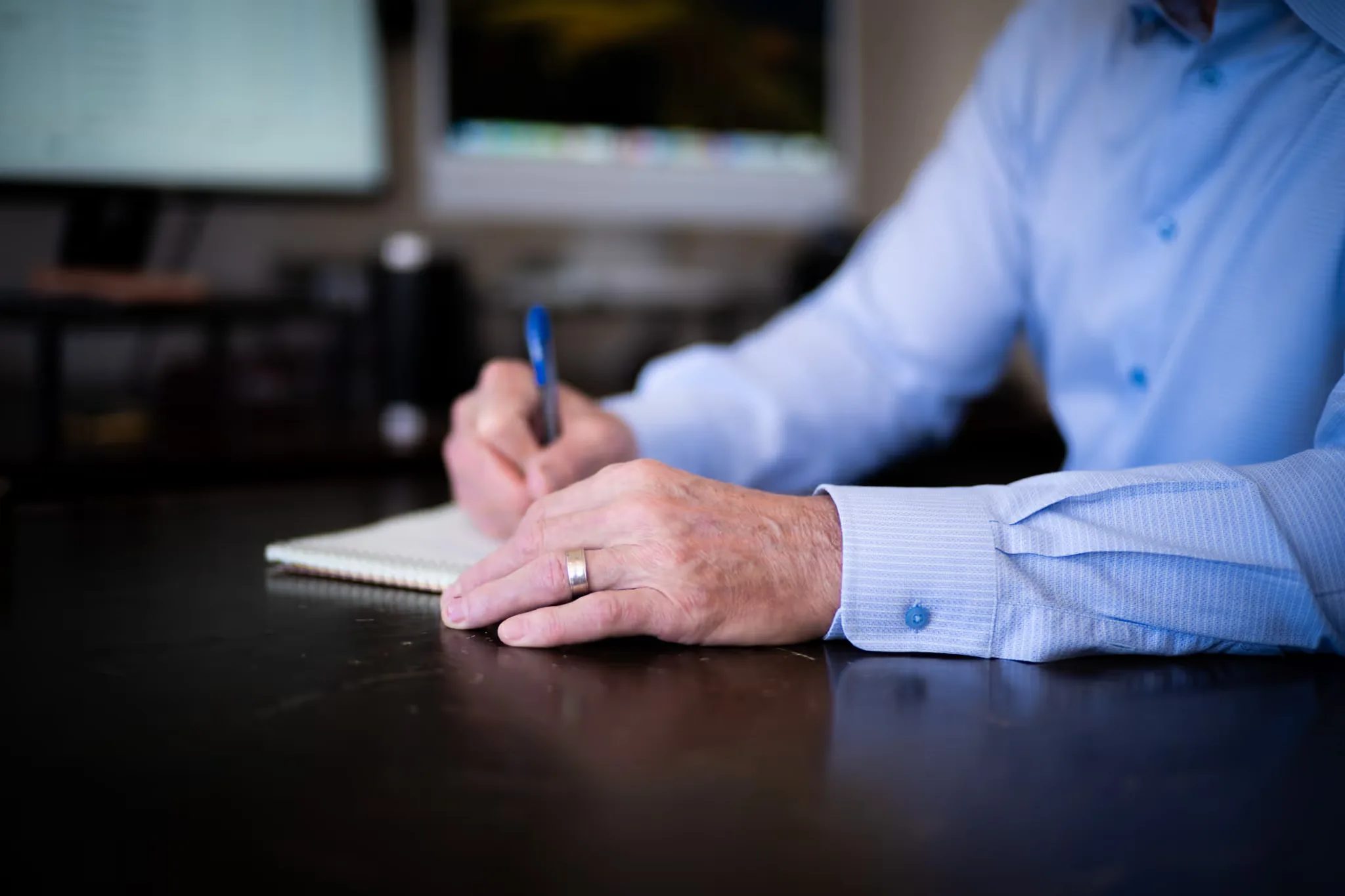 Close-up of hands writing on a notepad at a dark wood desk at Nelson Law Firm, monitors softly blurred behind.